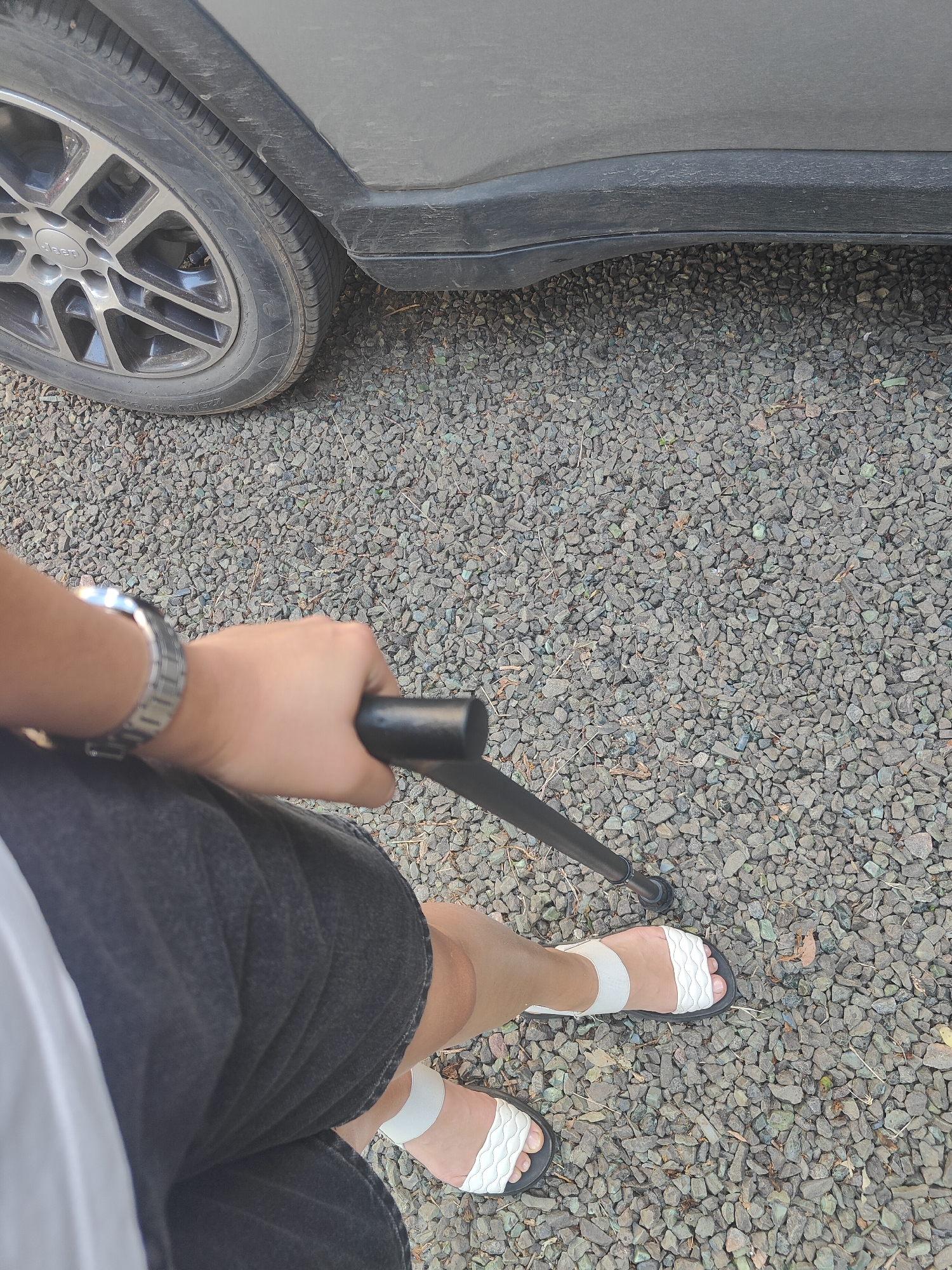A person is holding a black cane with their right hand, which is wearing a silver wristwatch. They are wearing dark shorts and white sandals. The person is standing on a gravel surface next to a dark-colored vehicle. The scene appears to be outdoors, possibly in Kryvyi Rih, Ukraine.