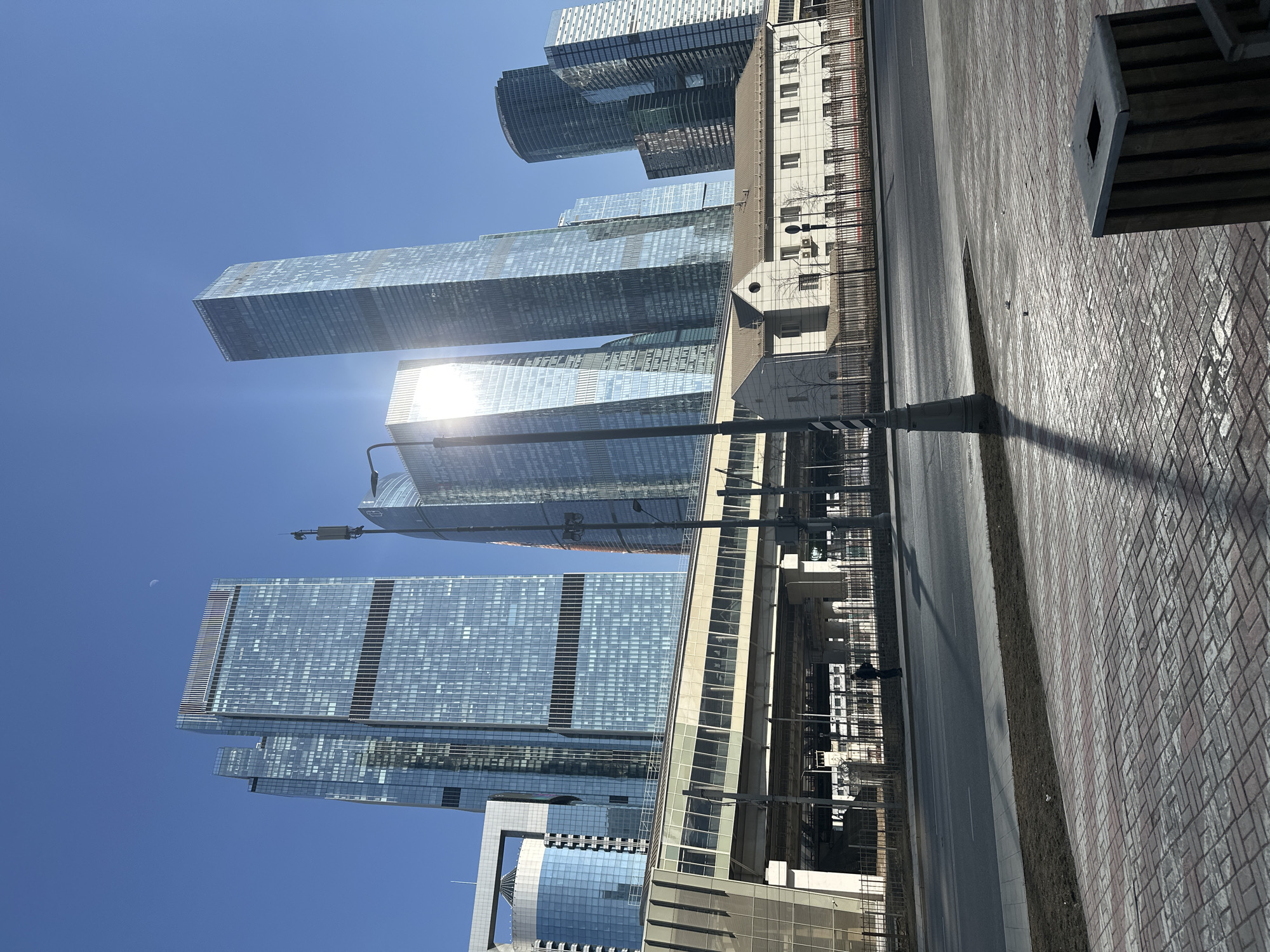 The scene is located in the city of Khimki, Russia, depicting a modern urban landscape under a clear blue sky. In the background, a cluster of multiple towering glass-clad skyscrapers dominates the skyline, their reflective surfaces catching bright sunlight. A strong sun glare emanates from a central skyscraper, obscuring some details. One prominent skyscraper features a dark horizontal band mid-section, while another on the far left incorporates an arch-like structural element near its top. A faint crescent moon is visible in the upper left portion of the sky.
In the mid-ground, an elevated multi-lane road or viaduct with prominent concrete support pillars spans horizontally across the frame. Integrated with this elevated structure is a modern pedestrian walkway or bridge, characterized by numerous horizontal windows. Beneath and slightly behind the elevated infrastructure, a two-story, older-style building with a light-colored facade, multiple rectangular windows, and a brown tiled roof is situated, providing a architectural contrast.
The foreground presents a wide, empty asphalt road, bordered by a strip of dry, sparse grass. To the right of the road, a brick-paved sidewalk with red and grey interlocking pavers extends towards the viewer. A single pedestrian, dressed in dark clothing, is walking on a paved area beneath the elevated structure, to the left of the older building. A tall, dark street light pole stands prominently in the mid-ground on the paved sidewalk, casting a long shadow towards the left. In the bottom right corner, a dark, rectangular trash receptacle with vertical slats and a metal top is visible on the sidewalk.