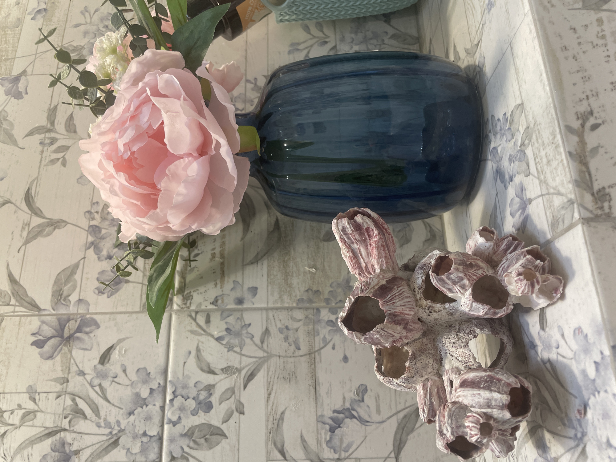 This is a close-up, eye-level, medium shot of a decorative arrangement on a surface that appears to be a shelf or countertop. The background consists of a wall tiled with a pattern of grey and blue floral motifs over a wood-grain texture.
In the foreground, to the left, is a cluster of barnacle-like shells, predominantly white with reddish-pink stripes. These shells are rough in texture and have hollow openings.
To the right of the shells is a deep blue, ribbed glass vase containing a large, artificial pink peony. Several small green leaves and sprigs of greenery accompany the peony. The vase is partially obscured by the peony. Behind the vase, a portion of a light blue, knitted object and a dark bottle are visible.
The surface the objects rest on is also tiled, with a similar floral pattern to the wall, but with lighter colors and a more prominent wood-grain effect. The lighting is soft and appears to be from the front, casting subtle shadows. There are no people or animals present in the image. The scene depicts a still life arrangement, likely indoors, in what could be a bathroom or decorative shelf. The provided location information is Taganrog, Russia.