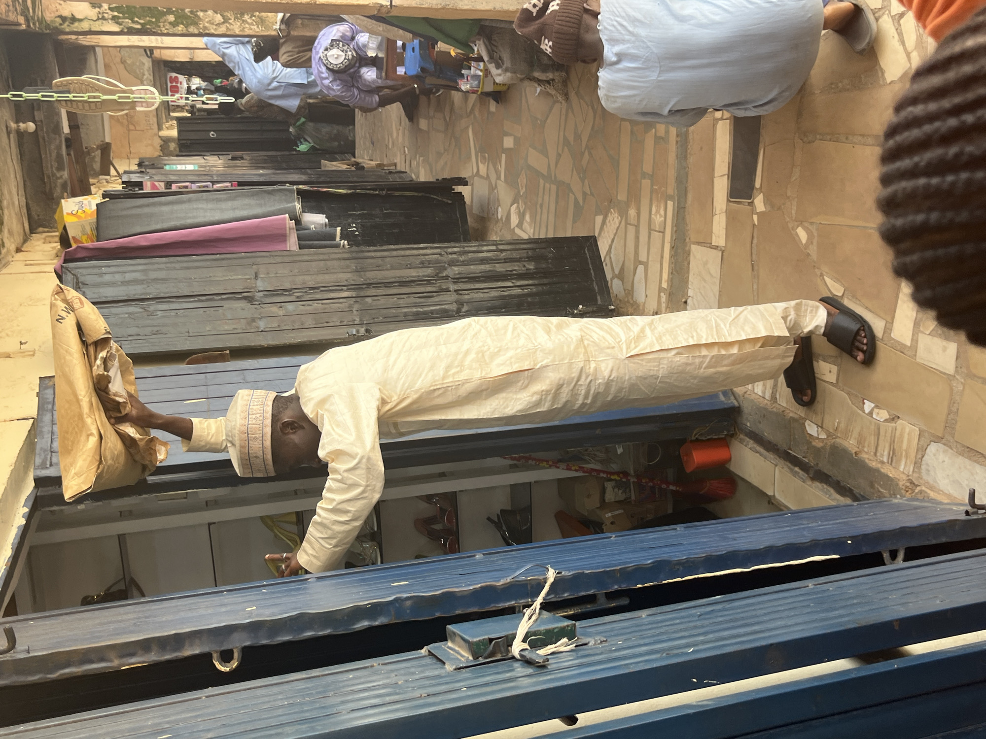 In the city of Jos, Nigeria, a man wearing a light-colored traditional attire and a cap is standing inside a shop, holding a bundle wrapped in brown paper over his head. The shop appears to be a retail establishment, with shelves visible behind him containing shoes. In the background, two other individuals are present; one is wearing a light blue garment and facing away from the camera, while another person is wearing a purple garment and appears to be kneeling or praying. The floor of the shop is tiled with a pattern of light and dark rectangular tiles. The shop's entrance is marked by a blue metal shutter that is partially open. The overall environment suggests an indoor marketplace or a commercial street.