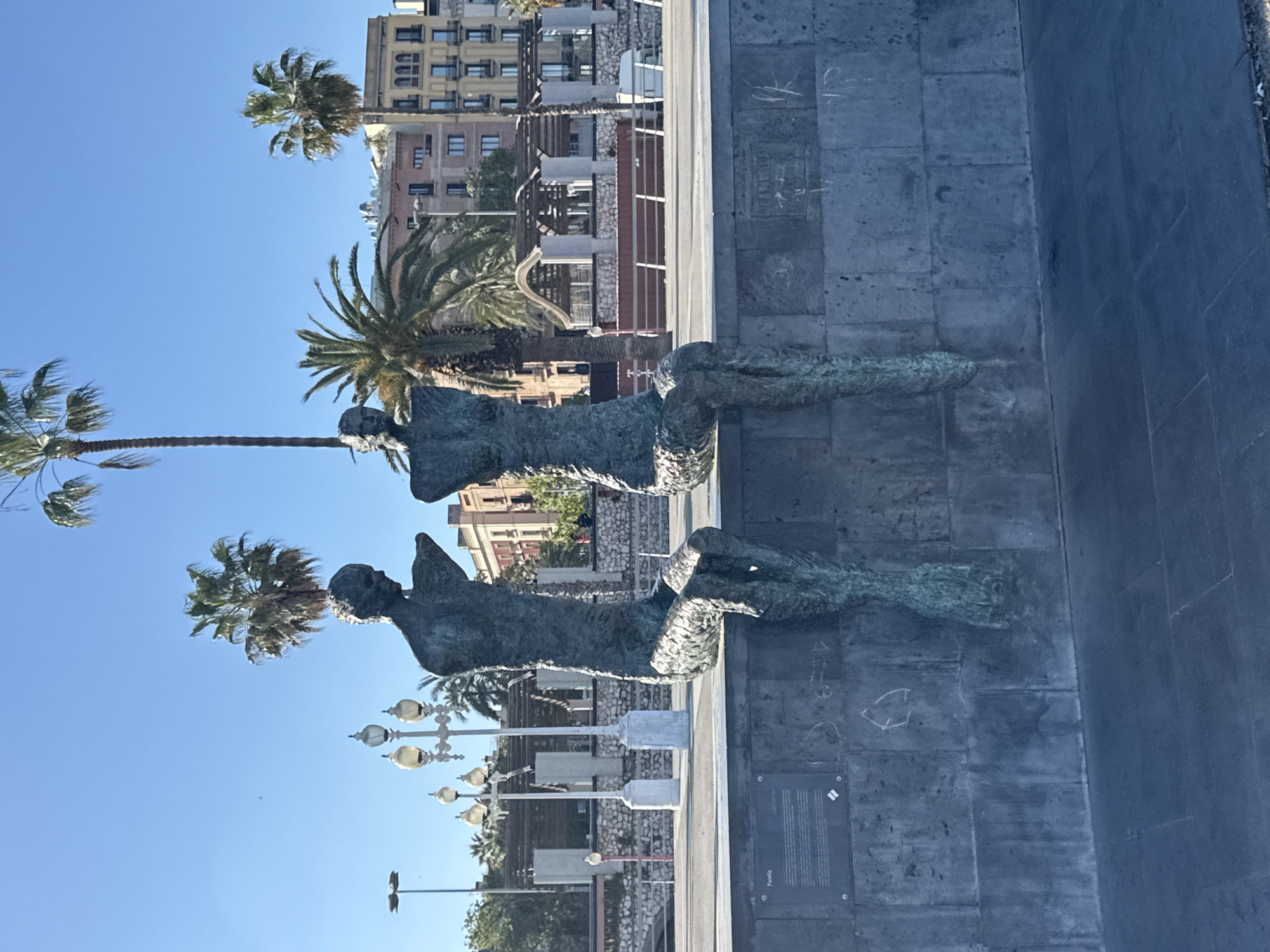 The image depicts the monument "A los Castellers" in Barcelona, Spain, photographed from a low angle on a clear day. The monument consists of two bronze figures, likely representing castellers involved in the construction of a human tower. In the background, palm trees line a street, with buildings visible further back. The monument rests on a stone base, which bears markings and what appears to be a plaque. The paving stones that surround the memorial have some graffiti.