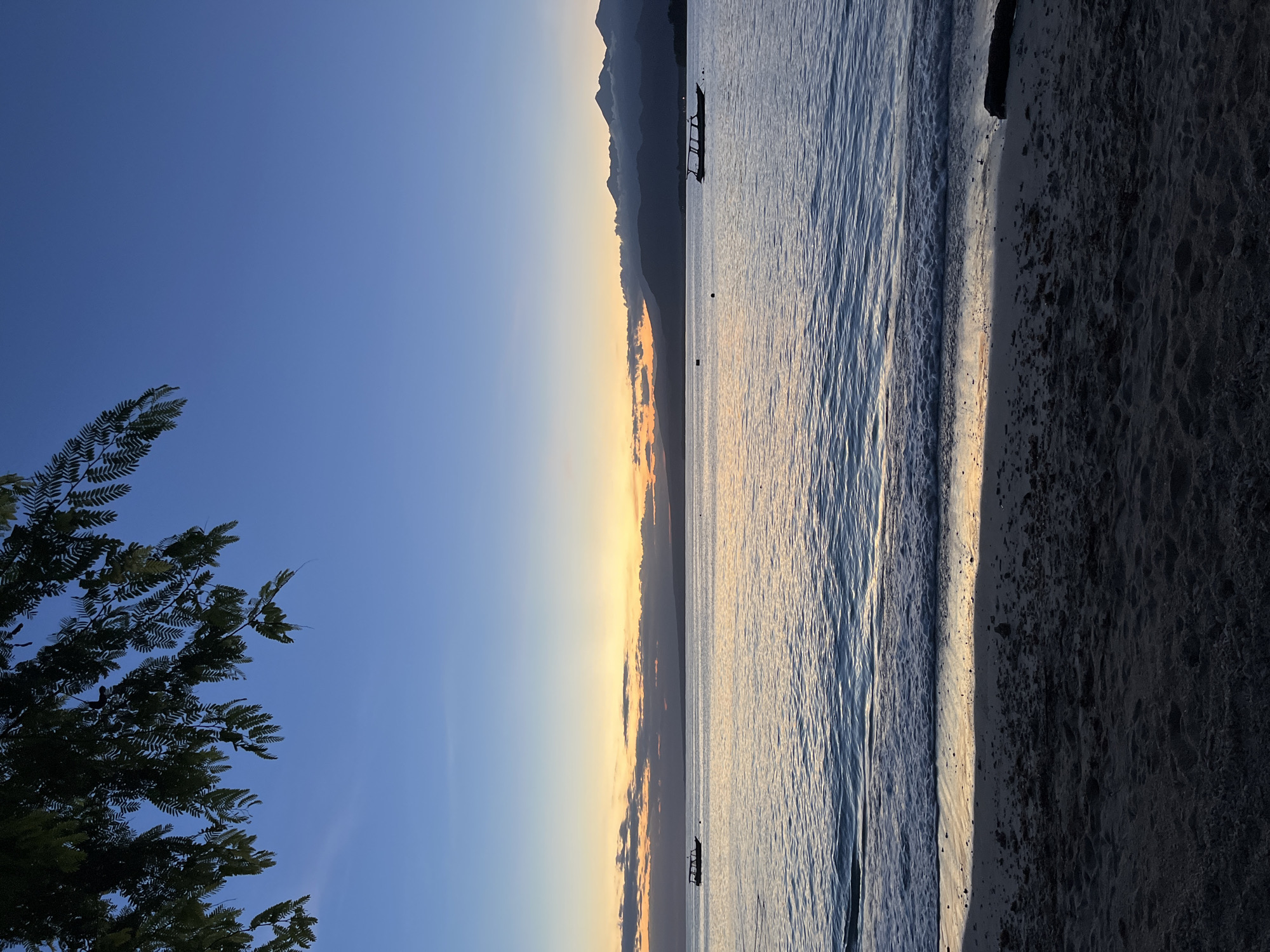 The image shows a beach scene at sunrise or sunset in Unknown, Indonesia. The sky is a gradient of blue to orange. Silhouetted mountains are visible on the horizon. Several boats are floating in the water. The water is calm with gentle ripples. The beach is sandy, with the sand near the water reflecting the light. A tree branch with leaves is in the bottom left of the frame.