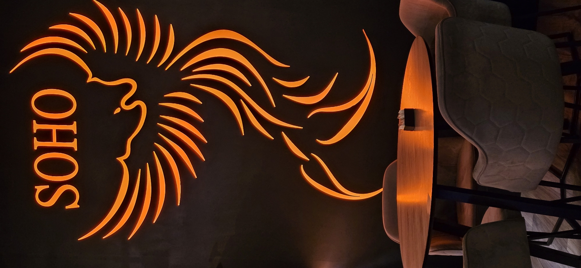 A dimly lit interior shot shows the word "SOHO" illuminated in orange neon script vertically on a dark wall, next to a stylized orange neon design resembling a bird or phoenix. To the right, there are parts of chairs and tables in muted neutral tones. Based on the Soho Caffe Bar, the location is likely Bar, Montenegro.