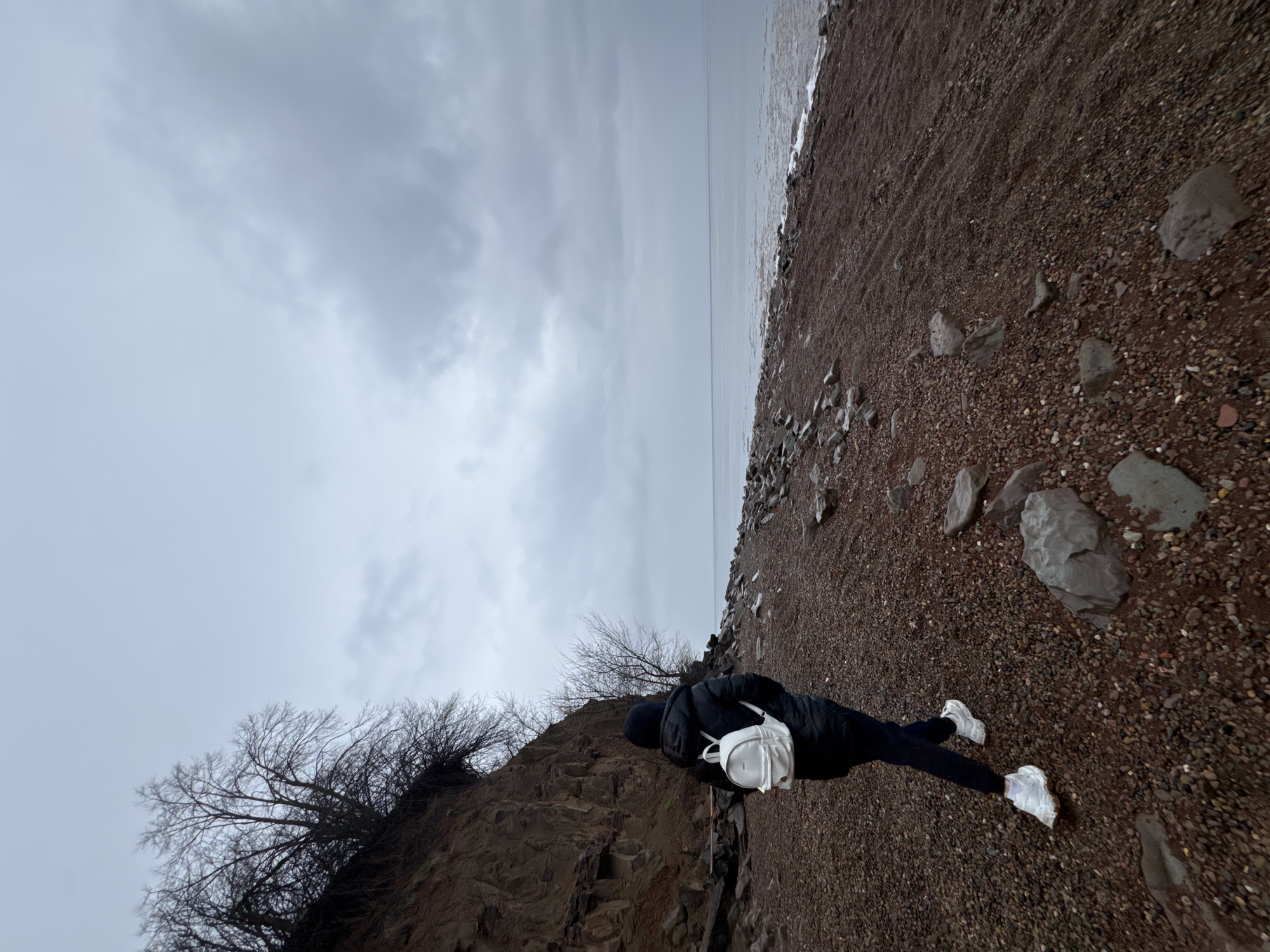 A person wearing a black jacket, dark pants, and white sneakers is standing on a red gravelly beach next to a body of water under a cloudy sky. The person has a white backpack. To the left of the person is a rocky cliff with bare trees. The location is outdoors and appears to be near the city of Kostomuksha in Russia based on the image's GPS coordinates.