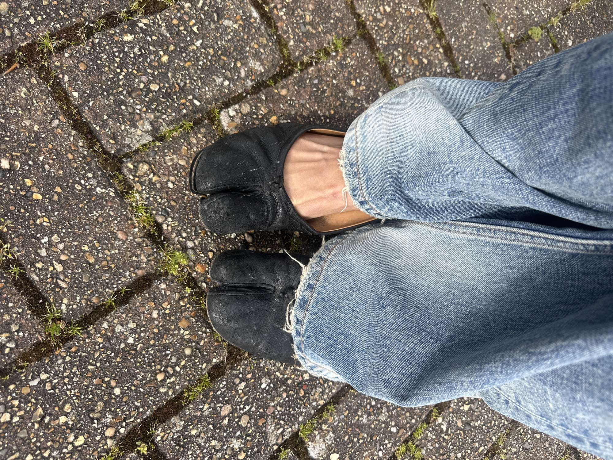 The image shows a person's legs from the knees down, wearing blue jeans and black split-toe shoes (Tabil shoes). The person is standing on a paved surface made of small stones and concrete with weeds growing in the cracks. The photo appears to be taken in Wealden, United Kingdom, based on the paving style and likely presence of weeds common to that region.