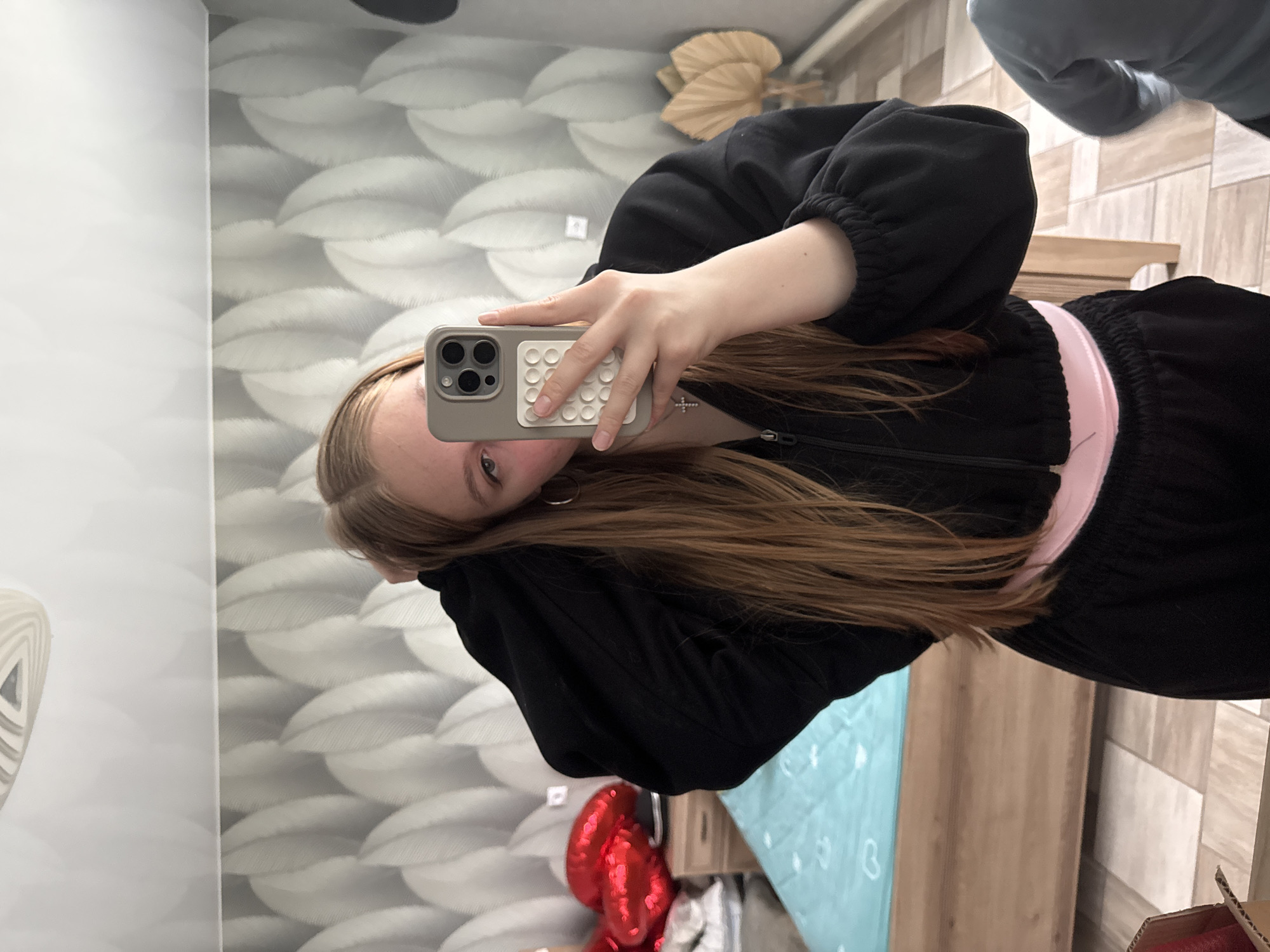 A young woman is taking a selfie in what appears to be a bedroom. She is wearing a black tracksuit with a pink waistband, and she has long, light brown hair. She is holding a smartphone with a gray case that has a grip on the back. A silver cross is visible on her neck.
The background includes a textured wall with a leaf pattern, a wooden headboard, and a light blue duvet on a bed. A small bedside table with red heart-shaped balloons is also visible. The exact location in Zhigalki, Russia, cannot be determined from the image.
