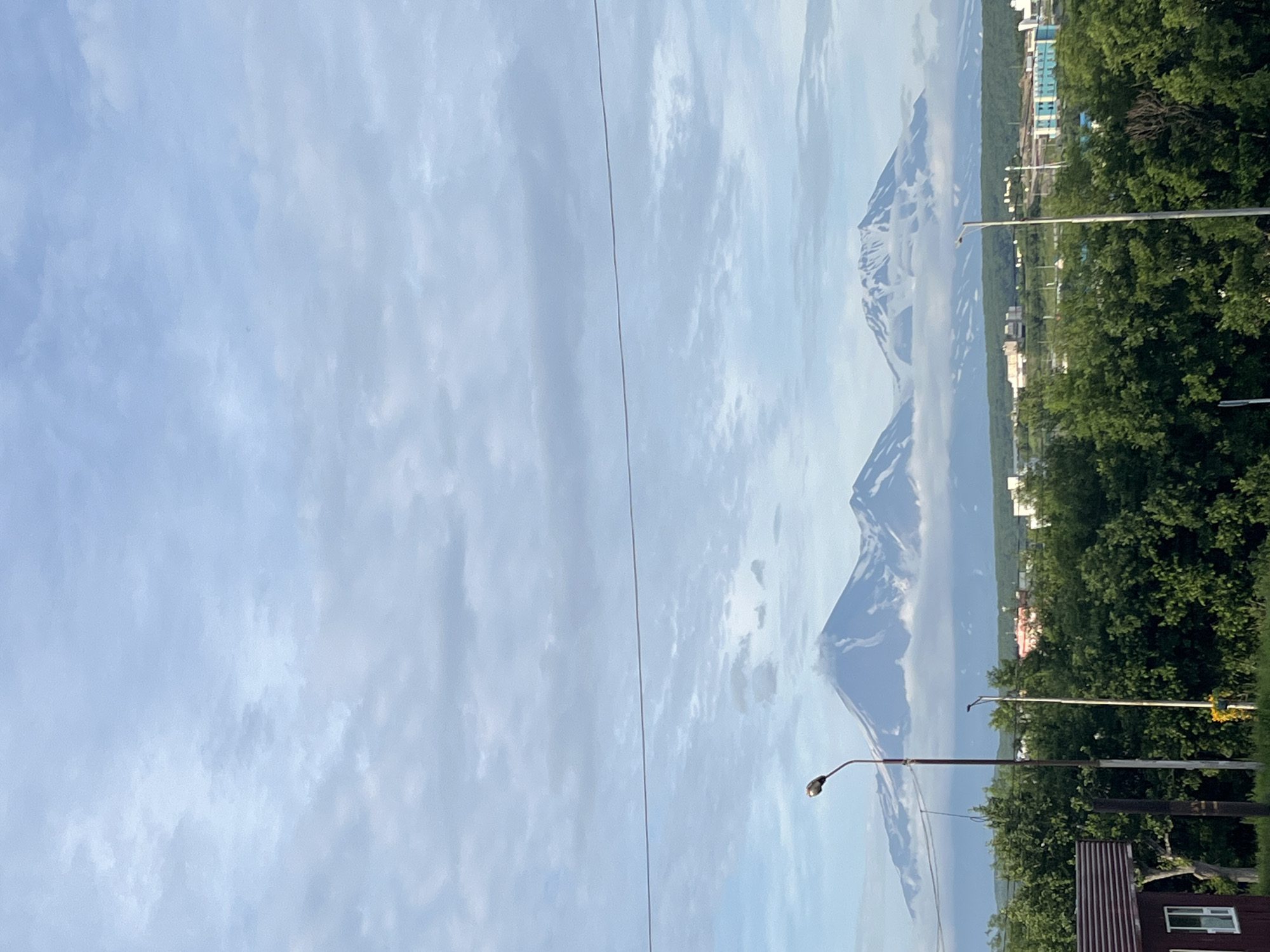 The image depicts a scenic view of the Avachinsky and Koryaksky volcanoes, iconic natural landmarks located near Petropavlovsk-Kamchatsky, Russia. In the foreground, lush green trees form a dense canopy, partially obscuring buildings and infrastructure. A tall, dark lamppost stands prominently on the left, with another lamppost visible to its right. A single horizontal wire stretches across the frame in the mid-ground. The imposing volcanoes, capped with snow, rise majestically in the background, their peaks partially veiled by clouds. A small cluster of buildings, some with light-colored facades and one with blue accents, can be seen nestled amongst the trees in the middle distance. The sky above is a soft blue, adorned with scattered, wispy clouds. The overall impression is one of a tranquil, early morning or late afternoon scene, showcasing the natural beauty surrounding the city.