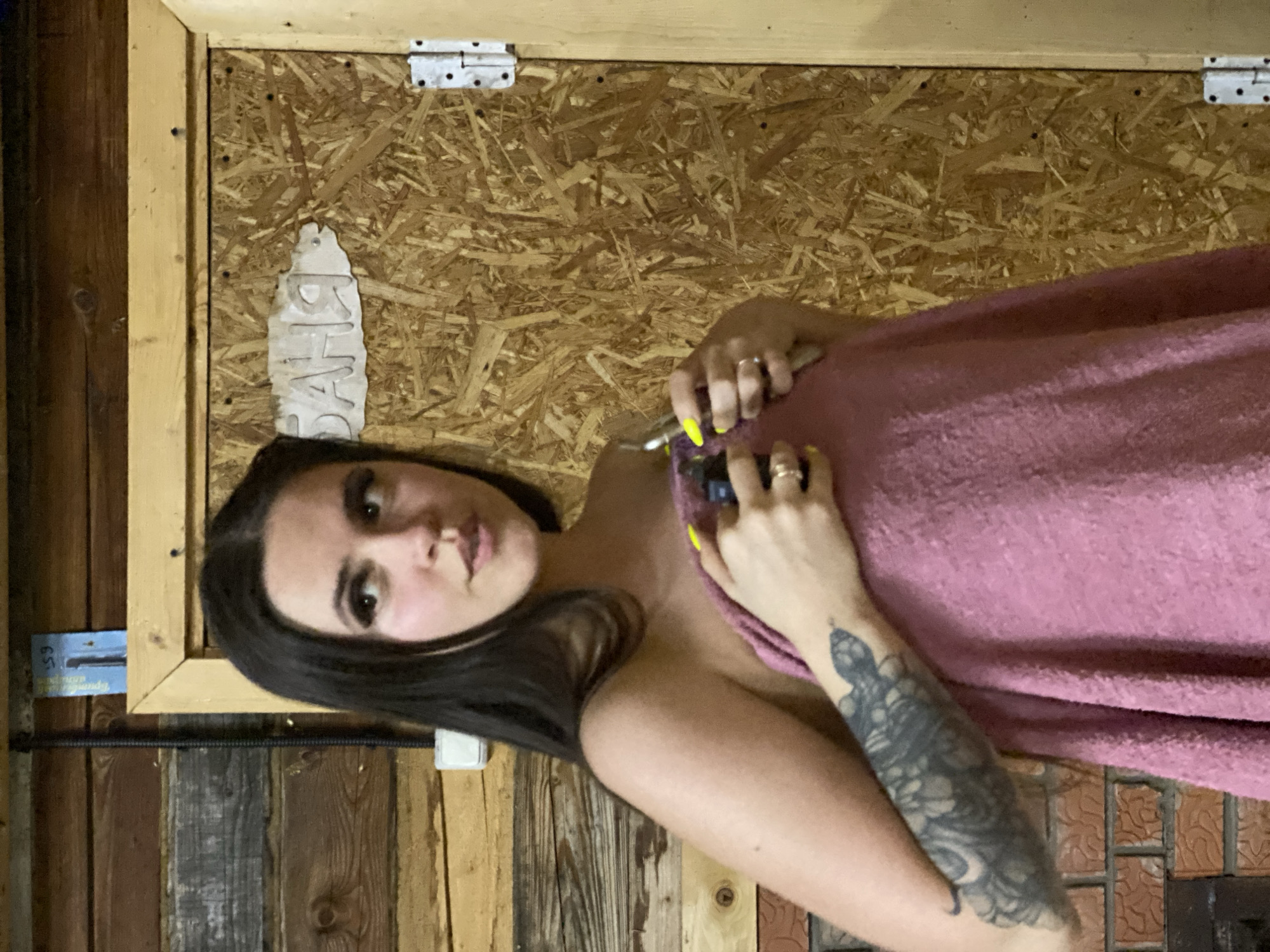 A woman with long dark hair and a tattoo on her left arm holds a pink towel. She is positioned in front of a wooden background with various textures and colors of wood. Above her, a wooden frame surrounds what appears to be particleboard. To the left, a blue package or label is partially visible. The location is indiscernible based on the visual information provided.