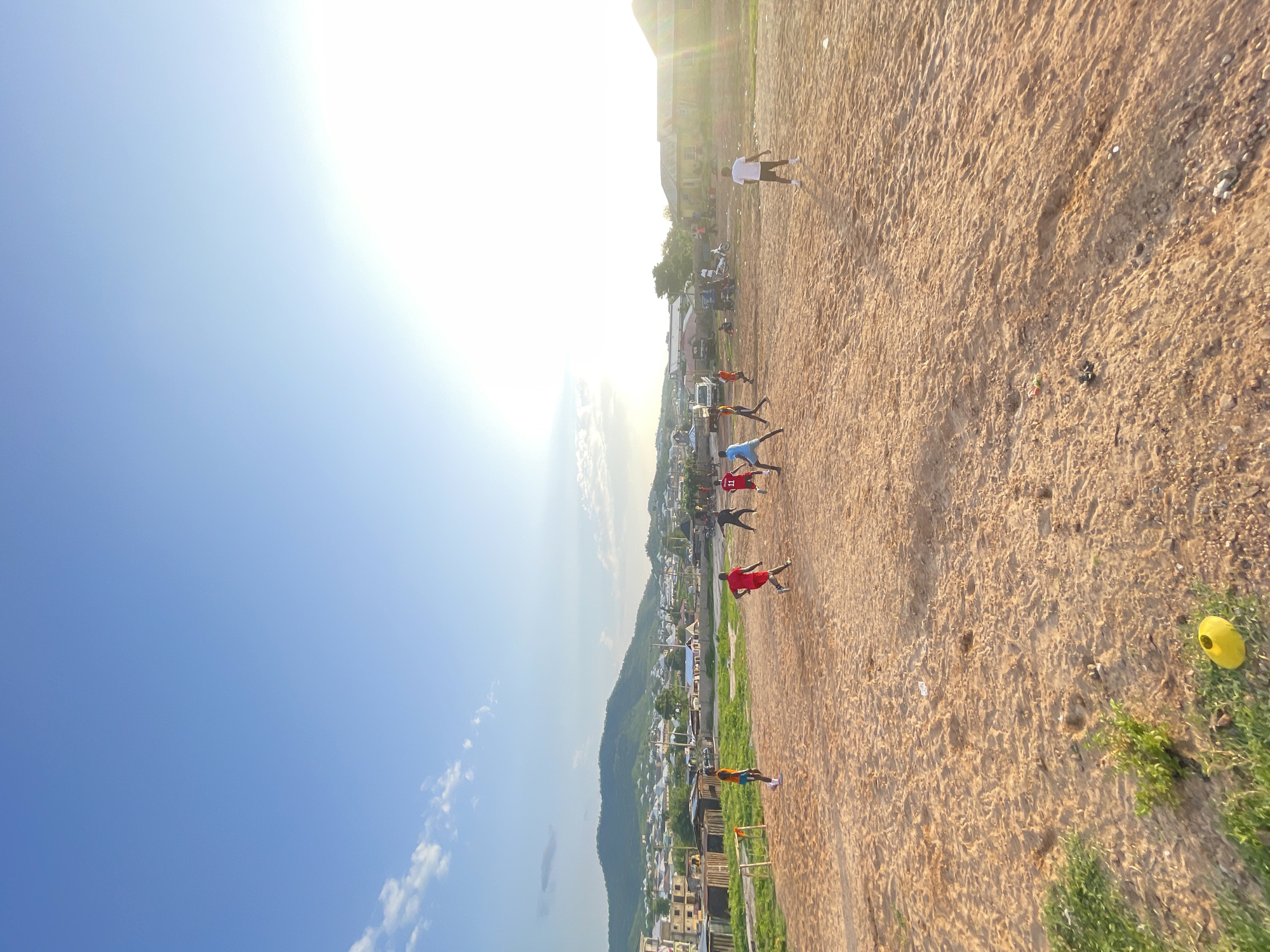 The image depicts a group of young men playing soccer on a dirt field in Lokoja, Nigeria. The sun is shining brightly, suggesting it is midday or late afternoon. The sky is a clear blue with some wispy clouds. The field is dusty and uneven, with scattered rocks and patches of dry grass. In the background, there are buildings and a lush green hillside, characteristic of a tropical or sub-tropical environment. The players are wearing casual clothing, mostly t-shirts and shorts, with some wearing jerseys. They appear to be actively engaged in the game, with some running and others in defensive positions. There is a yellow soccer cone on the grass near the right edge of the frame. The overall scene conveys a sense of youthful energy and camaraderie, with the players focused on their game despite the simple conditions of the playing field.