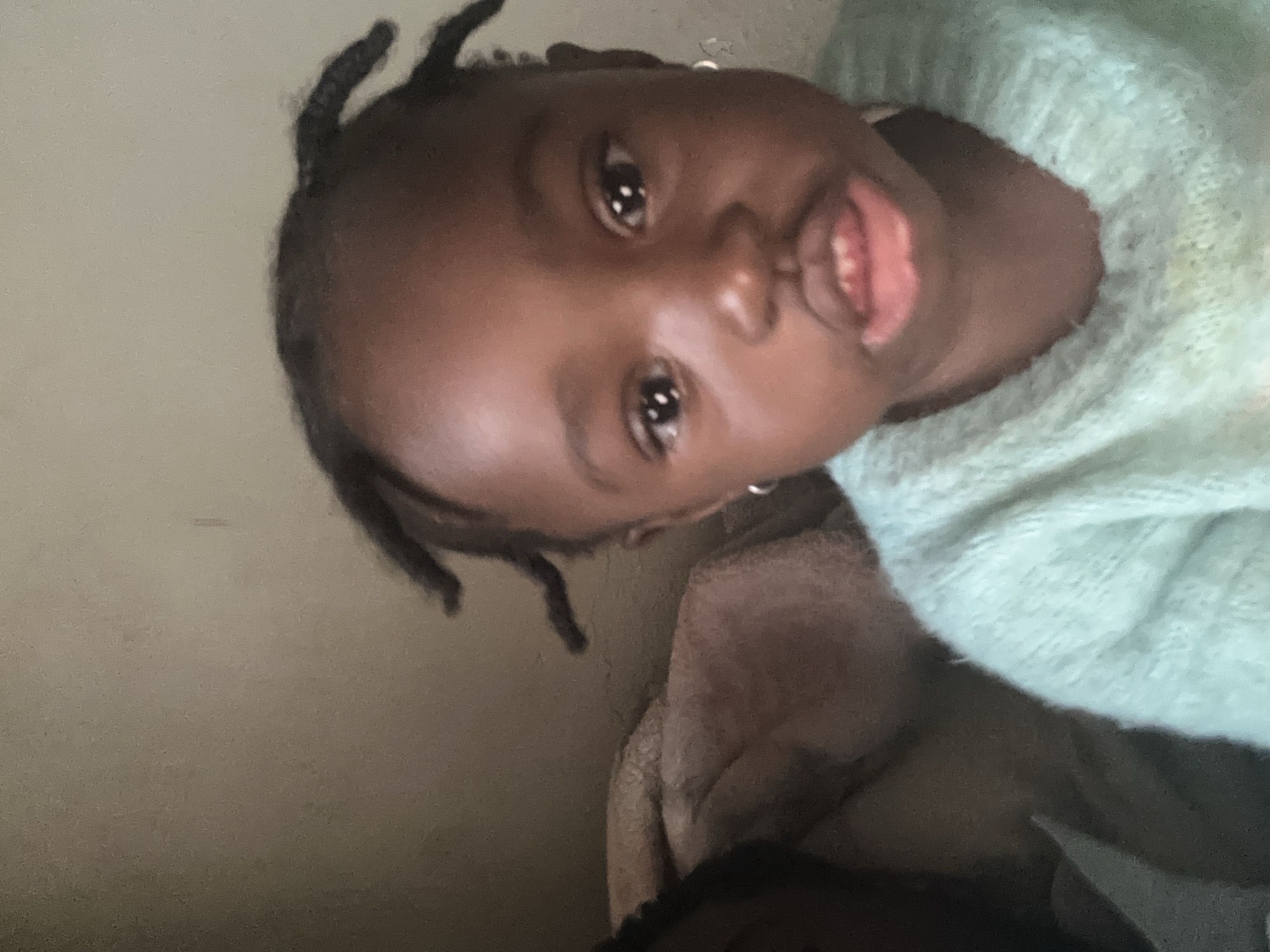An overhead shot captures a close-up of a young Black child's face, filling most of the frame. The child is wearing a light blue, fuzzy sweater. Their dark hair is styled in small braids with some loose ends. The child has large, dark eyes and their mouth is slightly open, revealing a hint of their tongue and lower teeth. A small hoop earring is visible on their right ear. In the background, a person's head and shoulders are visible on the left, partially obscured and out of focus, also appearing to be wearing a light-colored garment. The background itself is a plain, light-colored wall.
