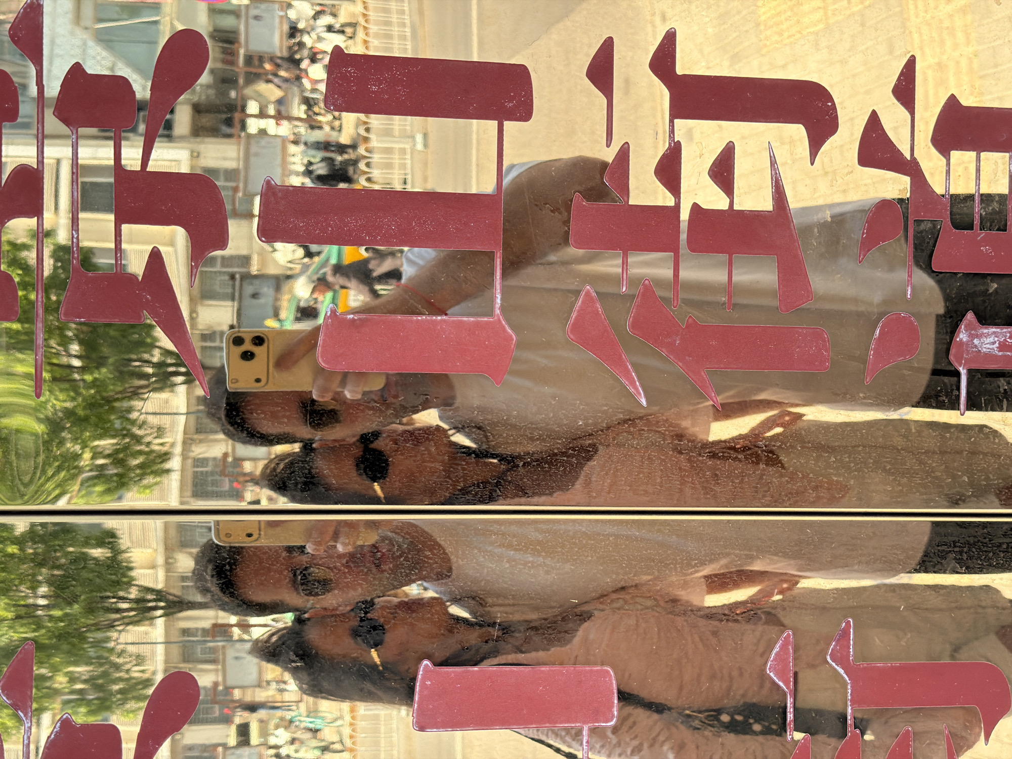 The image shows a person's reflection in a glass storefront in the Dongcheng District, China. The person is holding a phone, presumably taking a picture of themselves or the storefront. Red, stylized Chinese characters are affixed to the glass, partially obscuring the reflection. The reflection also includes a street scene with buildings, trees, and vehicles, suggesting a busy urban environment. The perspective is slightly angled, giving a dynamic feel to the scene.