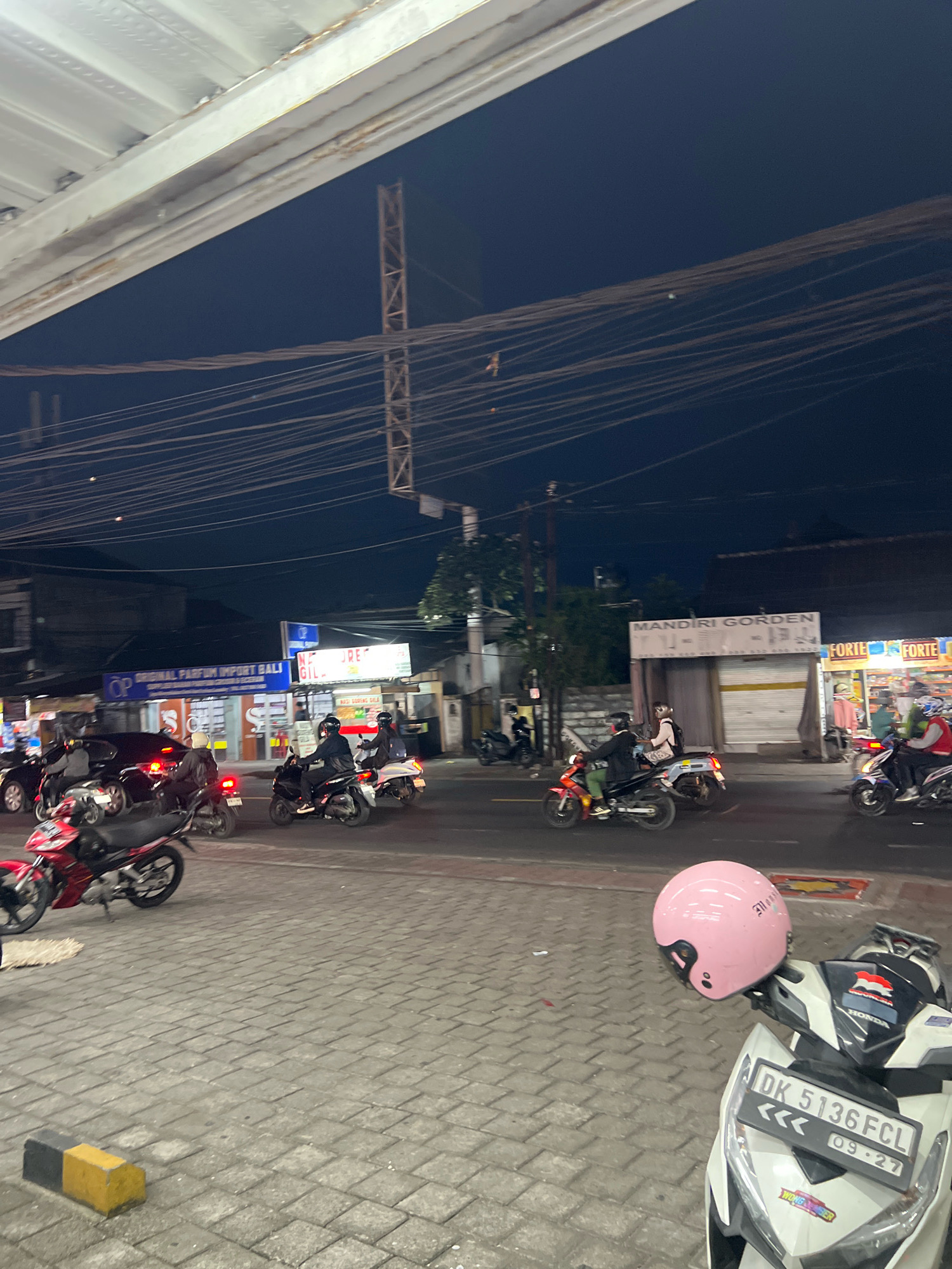 This is a nighttime street scene in Denpasar, Indonesia, with several motorcycles and scooters on a paved road. In the foreground, a white scooter with a pink helmet is parked. The scooter has a license plate that reads "DK 5136 FCL" and "09-27." Further down the road, other motorcycles are visible, some with riders. The buildings lining the street appear to be shops with illuminated signs. The sky is dark, indicating it is nighttime, and there are many overhead power lines crisscrossing the scene. The overall impression is one of a bustling urban environment at night, likely a commercial area.