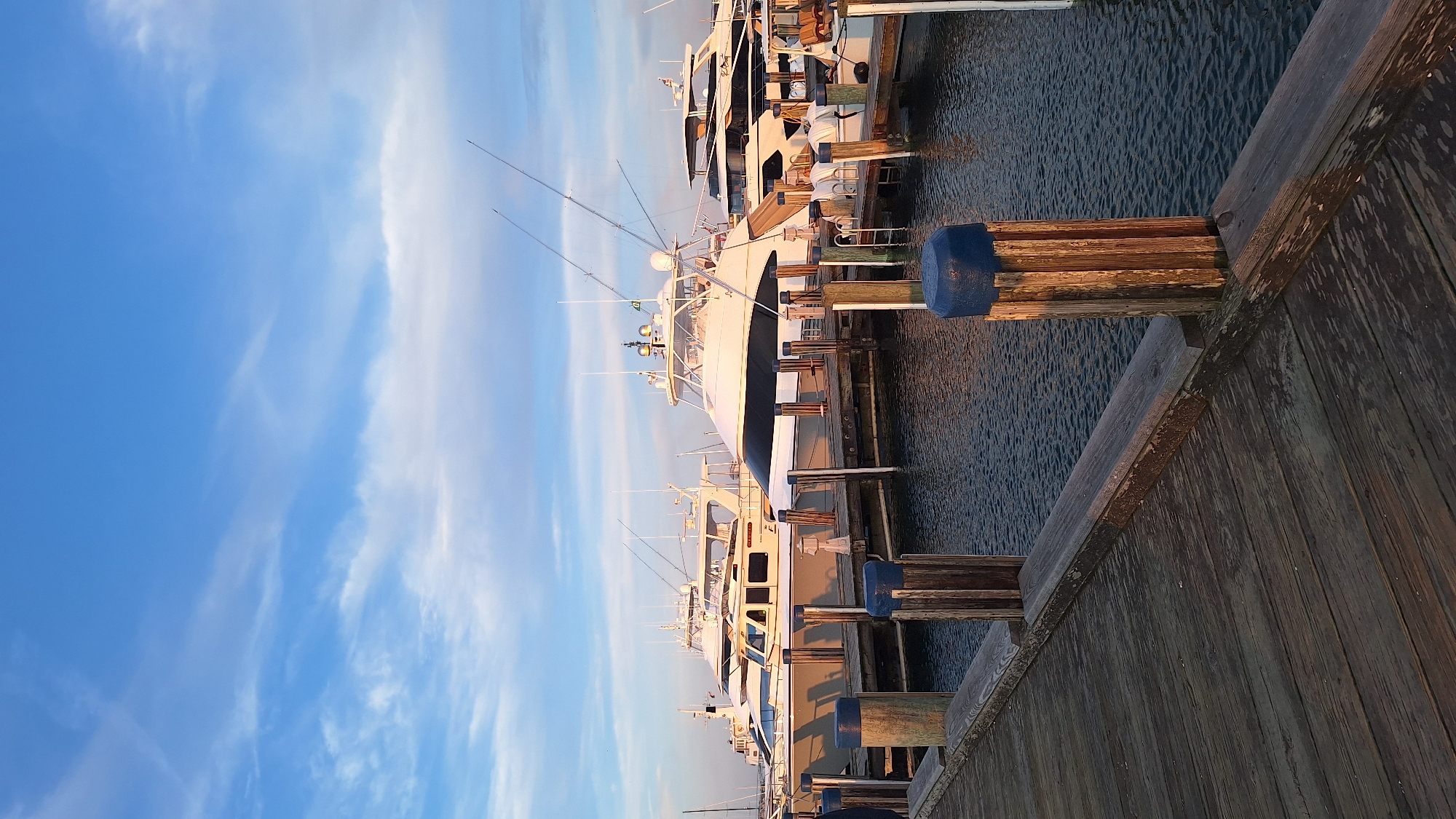 A vertical, eye-level shot captures a marina on Nantucket, Massachusetts, USA, at sunset. In the foreground, a wooden pier extends diagonally from the lower left corner into the frame, its weathered planks showing signs of age and exposure to the elements. Several wooden pilings topped with blue caps are spaced along the pier. The water of the marina, reflecting the sky, appears dark and rippled.
Dominating the mid-ground and background are several large, white yachts docked side-by-side. These vessels are modern and well-maintained, with various structures, antennas, and fishing rods visible on their decks and superstructures. The setting sun casts a warm, golden light on the scene, illuminating the boats and the sky. The sky itself is a clear blue with streaks of wispy white clouds, creating a serene and picturesque atmosphere. The overall impression is one of tranquility and nautical elegance.