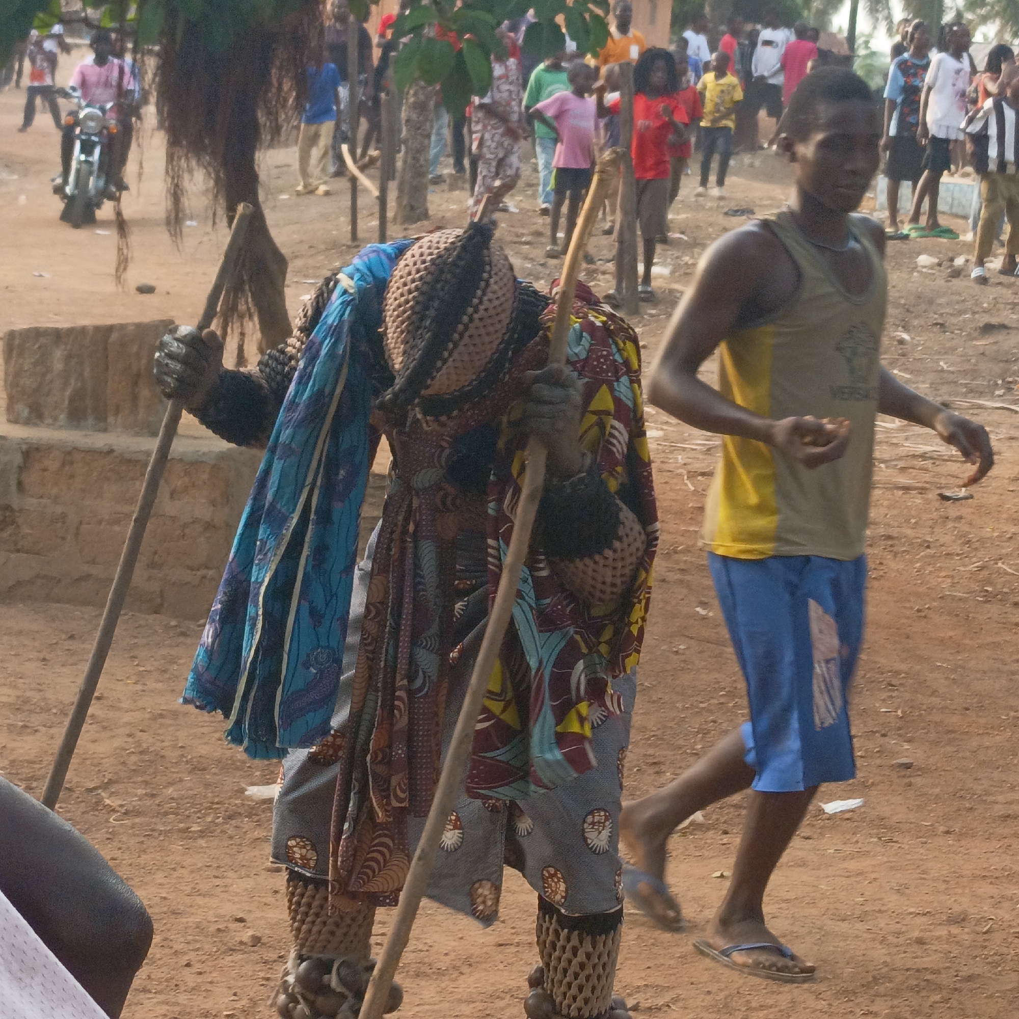 In Akwanga, Nigeria, a person dressed in a ceremonial costume stands holding two poles. The costume includes a woven mask, patterned clothing, and woven leggings with beaded accents. A man in a yellow tank top and blue shorts walks in front of the costumed person. In the background, a crowd of people and a motorcycle are visible. The ground appears to be dirt.