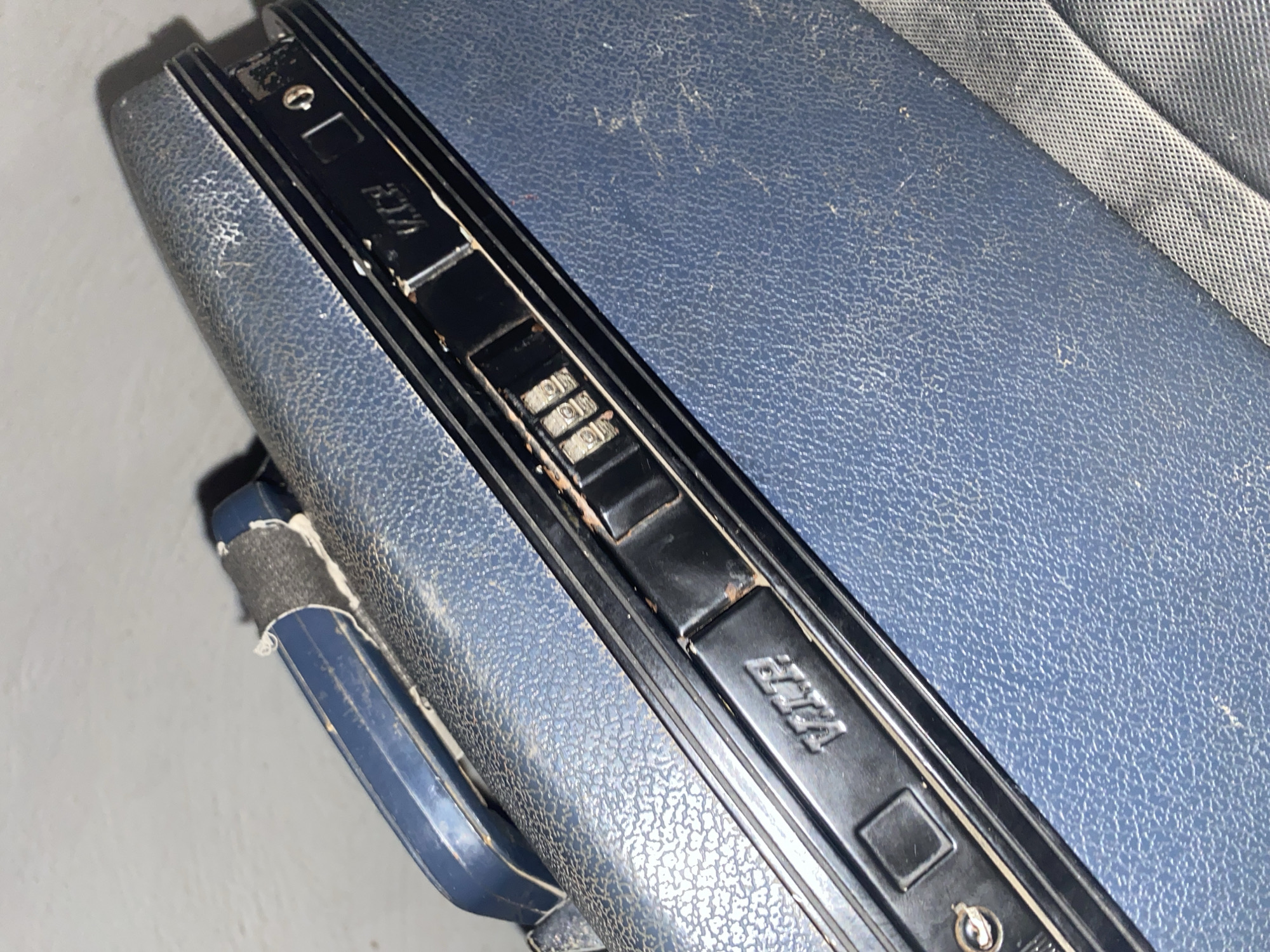 The image shows a close-up, angled view of a blue, textured suitcase. The suitcase is lying on a light-colored, slightly textured surface. Visible on the side of the suitcase are black plastic components, including a handle, a cassette player mechanism with visible buttons and dials, and lettering that appears to read "VIFA". The surface of the suitcase shows signs of wear and dust. A grey, woven strap is partially visible along the bottom right edge of the image, running diagonally across the suitcase. The lighting is somewhat dim, casting shadows and highlighting the textures of the suitcase and the surface it rests on.