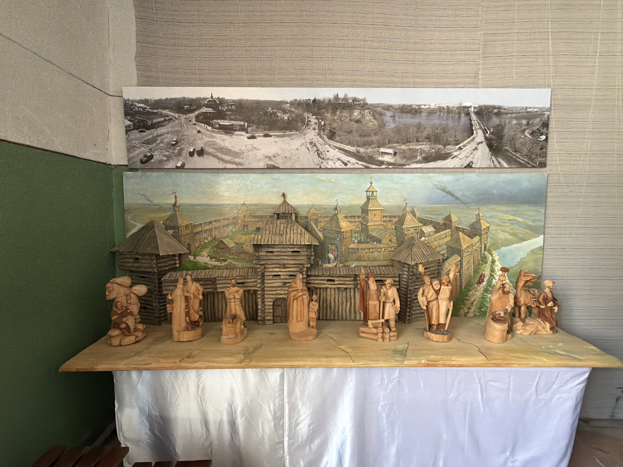 The image displays a diorama on a table covered with a white cloth. The diorama features a backdrop of two horizontal artworks. The upper artwork is a black and white photograph of a city landscape with a bridge. The lower artwork is a colorful painting of a wooden fortress. In front of the painted fortress, there are several small wooden figurines, depicting people and animals. The table is set against a wall with green and beige textured surfaces. The scene appears to be indoors, possibly in a museum or exhibition space.