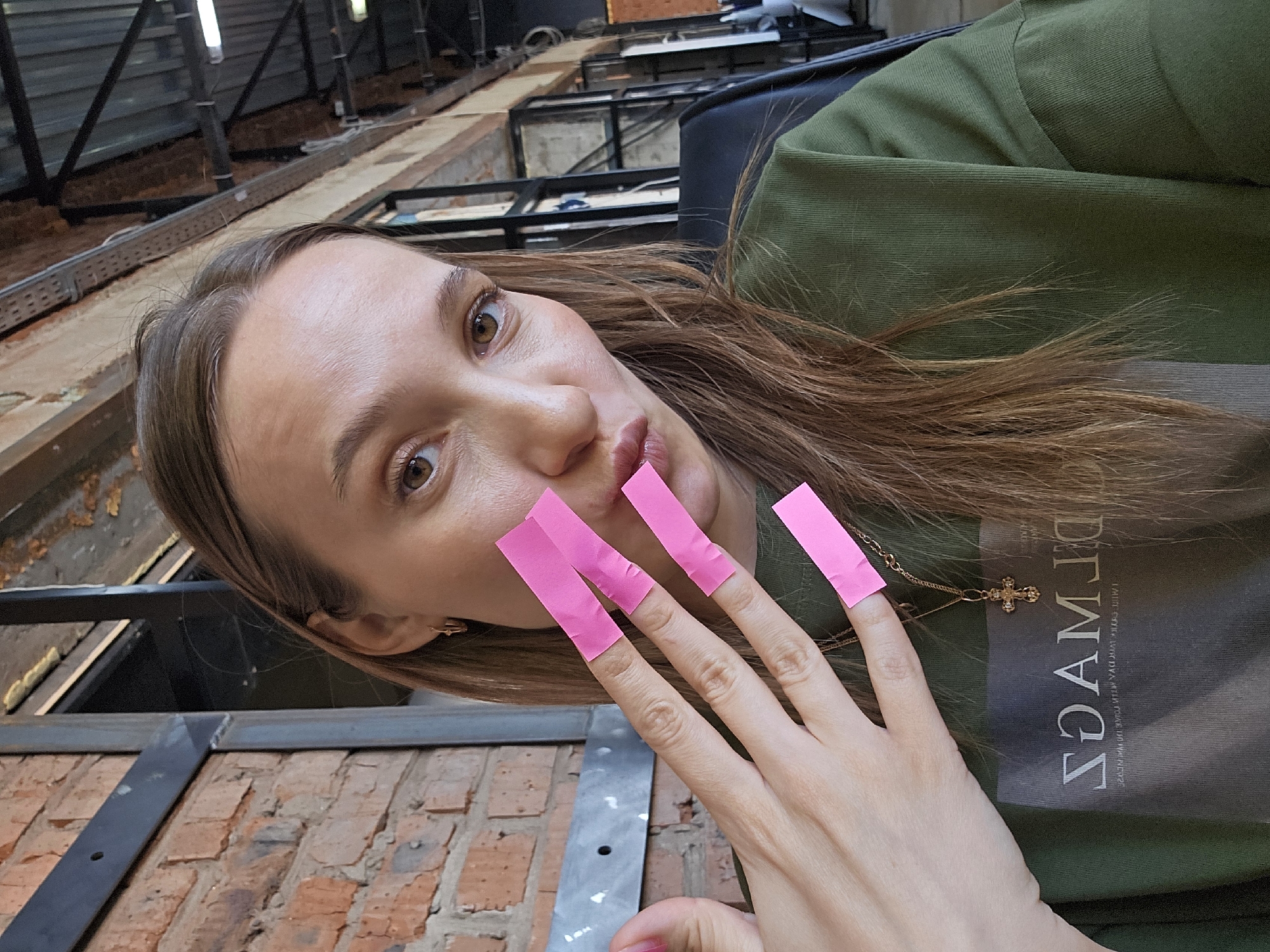 A selfie taken in Chaikovsky, Russia, shows a woman with light brown hair, fair skin, and brown eyes. She's wearing a green long-sleeved shirt with a gray section and a gold necklace with a cross pendant. Her fingers are adorned with pink rectangular strips. The background features an industrial-looking setting with exposed brick, metal structures, and overhead beams.