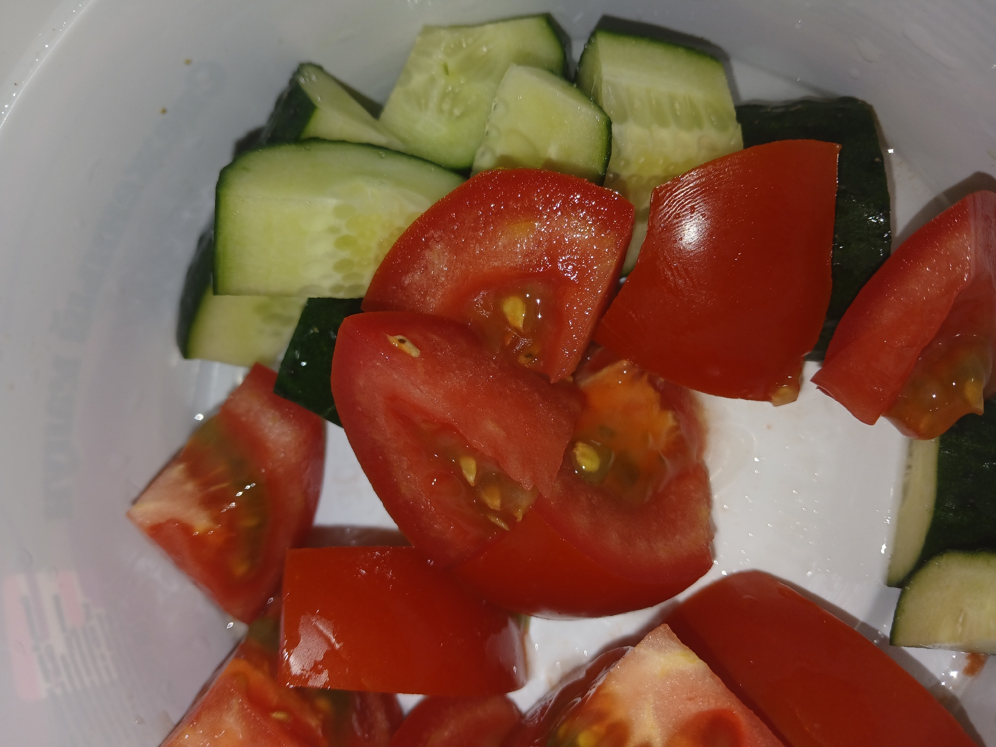 A close-up, overhead shot shows chopped tomatoes and cucumbers in a white bowl. The tomatoes are bright red and appear juicy, some with visible seeds. They are cut into wedges and irregular pieces. The cucumbers are sliced into rounds and then cut into smaller, mostly rectangular pieces. The outer skin of the cucumbers is dark green, while the inner flesh is pale green with a lighter, seedy core. The vegetables are wet, suggesting they have recently been washed or prepared for a meal. The white bowl is visible in the background and to the right, with a slightly textured surface. The lighting is bright, casting a slight sheen on the wet vegetables. The overall scene is one of fresh, prepared ingredients, likely for a salad or side dish. There is no visible text in the image.
