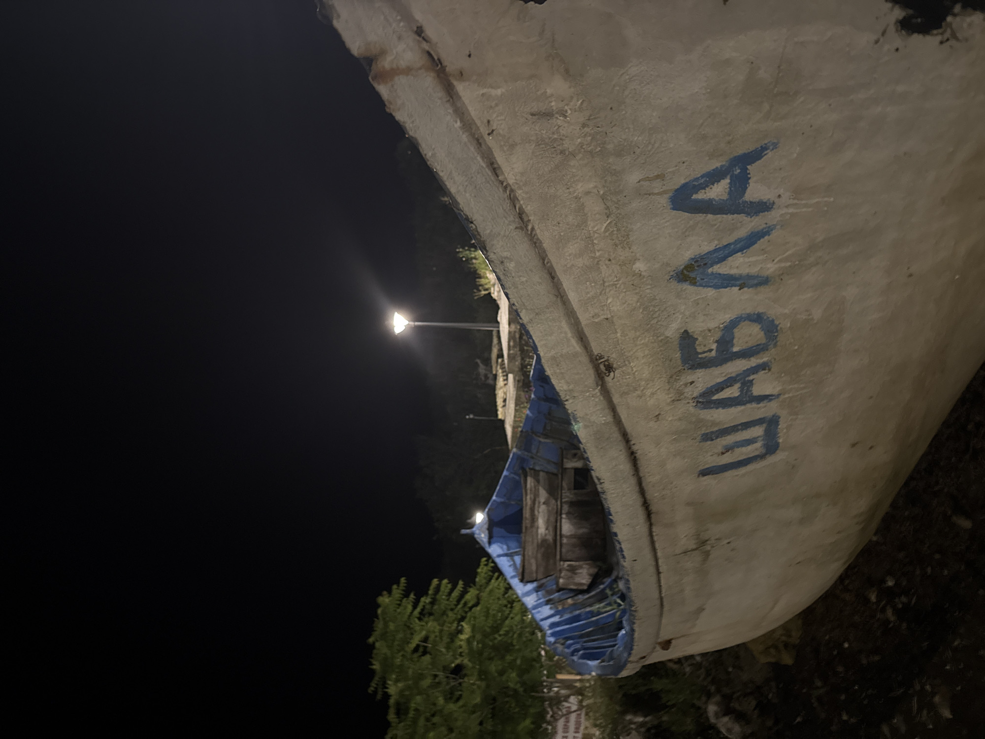 The image shows a close-up, low-angle view of the side of a white and blue boat at night, resting on land, with the name "ШАБЛА" (Shabla) painted in blue on its hull. The boat appears weathered and worn, with visible paint peeling and rust stains. The interior of the boat, visible at the front, shows blue paint and wooden seating. In the background, a modern streetlamp illuminates the scene, casting light on trees and vegetation. The sky is dark, indicating nighttime. The boat is located in the city of Tyulenovo, Bulgaria.