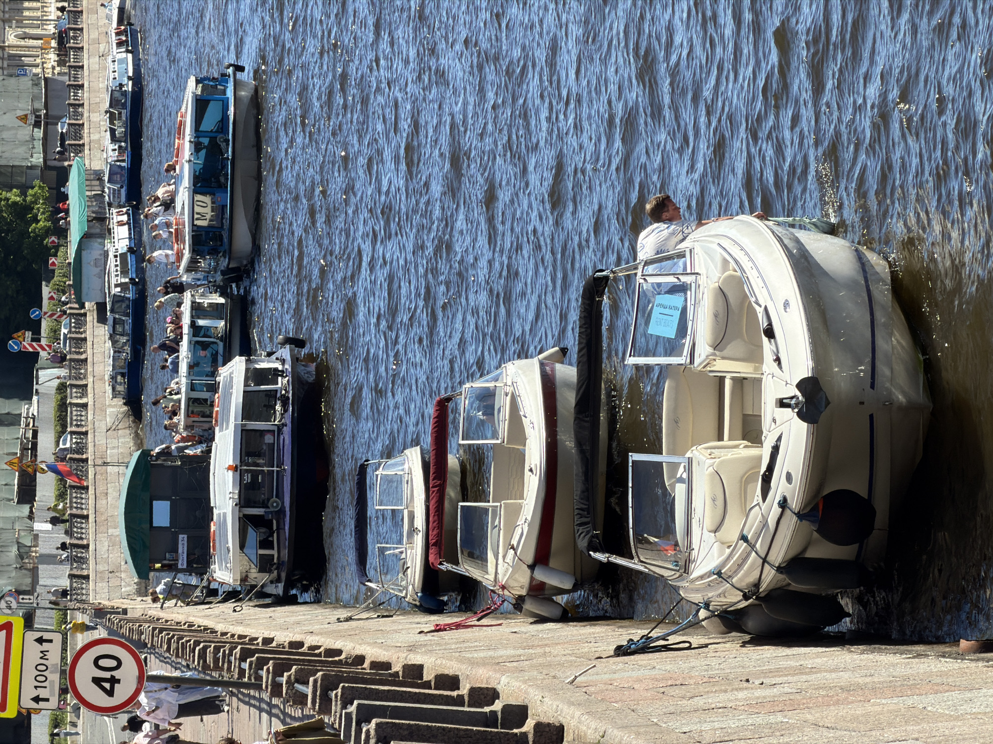 This image captures a vibrant, sunny daytime scene on an urban waterway, specifically identified as the Fontanka River in Saint Petersburg, Russia, by the visible text "ФОНТАНКА 34" on one of the boats and a Russian flag in the background.
In the foreground, a light-colored motorboat, possibly a Bayliner model, is moored parallel to a stone embankment on the left. A man in a light-patterned shirt is leaning over its starboard side, attending to something near the waterline. The boat features a folded-back black bimini top and a blue sign with white Cyrillic text, "АРЕНДА КАТЕРА" (Boat Rental), displayed on its windshield. Several black fenders cushion the boat against the rough-hewn stone embankment. Three additional similar, smaller motorboats are docked further along the embankment to the left, each equipped with fenders and featuring different colored bimini tops (maroon and dark blue/black). Mooring lines secure these boats to the embankment.
The embankment itself is constructed from large, light-colored stone blocks, forming a solid wall. Pedestrians are visible walking on the pavement above the embankment. A circular red-bordered "40" speed limit sign is prominently displayed on a pole, along with an arrow sign indicating "100M ↑".
Further out in the blue, rippling water, multiple larger passenger boats are visible. One prominent blue-hulled tour boat has "МО" in white Cyrillic letters on its cabin, and another behind it displays "ФОНТАНКА 34" on its stern. These boats are carrying groups of people, suggesting sightseeing activity. The water shows significant surface chop and glints of reflected sunlight.
In the distant background, across the waterway, an opposing embankment with more buildings and sparse foliage is visible. A Russian flag with white, blue, and red horizontal stripes flies from a pole on that distant bank. Numerous people are also discernible on the far embankment. The overall scene suggests a busy tourist area on a clear day.