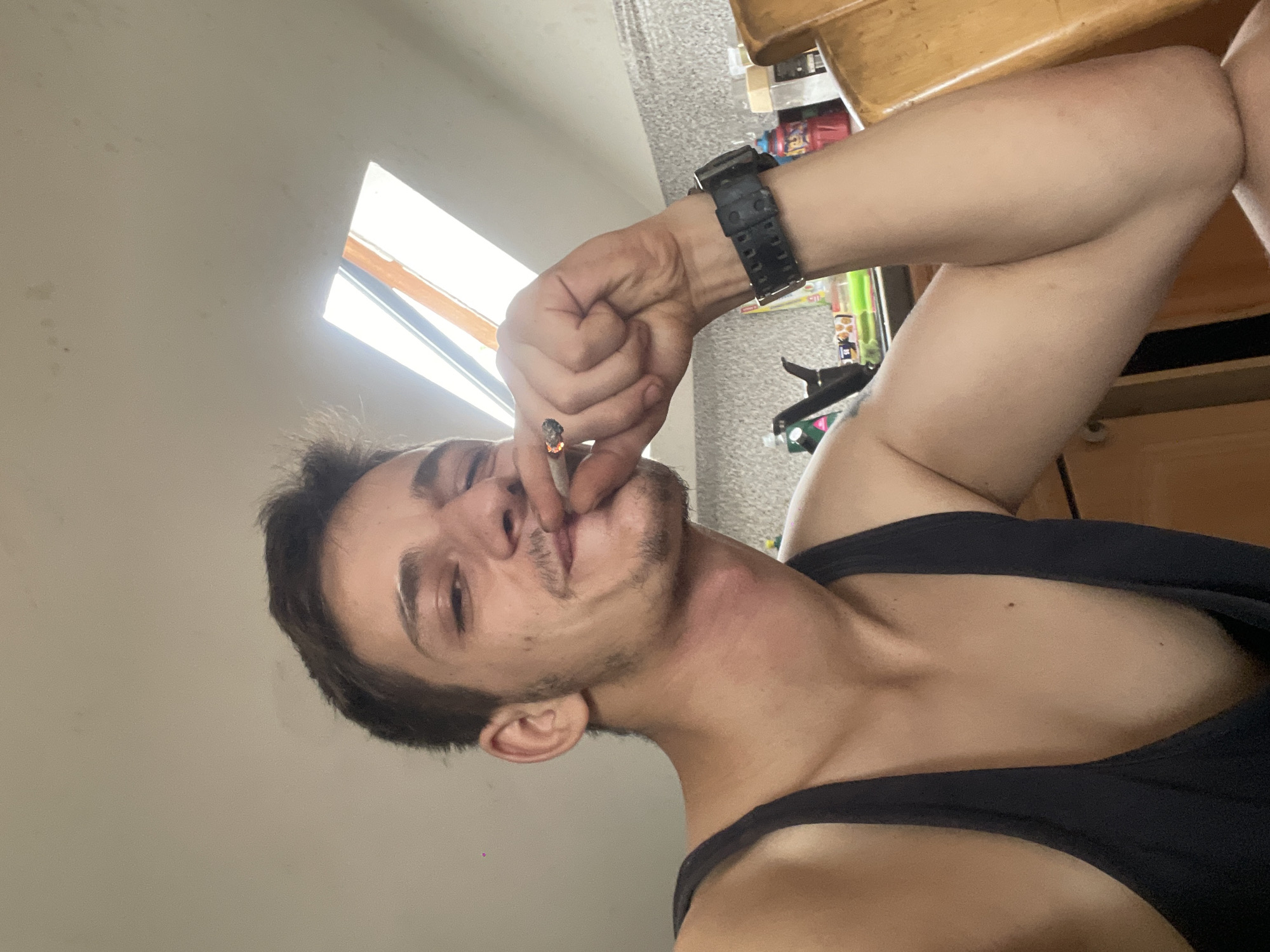A man is indoors, holding a cigarette to his lips and looking at the camera. He is wearing a black tank top and a black watch on his left wrist. The background is blurred but appears to be a kitchen area with cabinets and a skylight in the ceiling. The lighting suggests it is daytime.