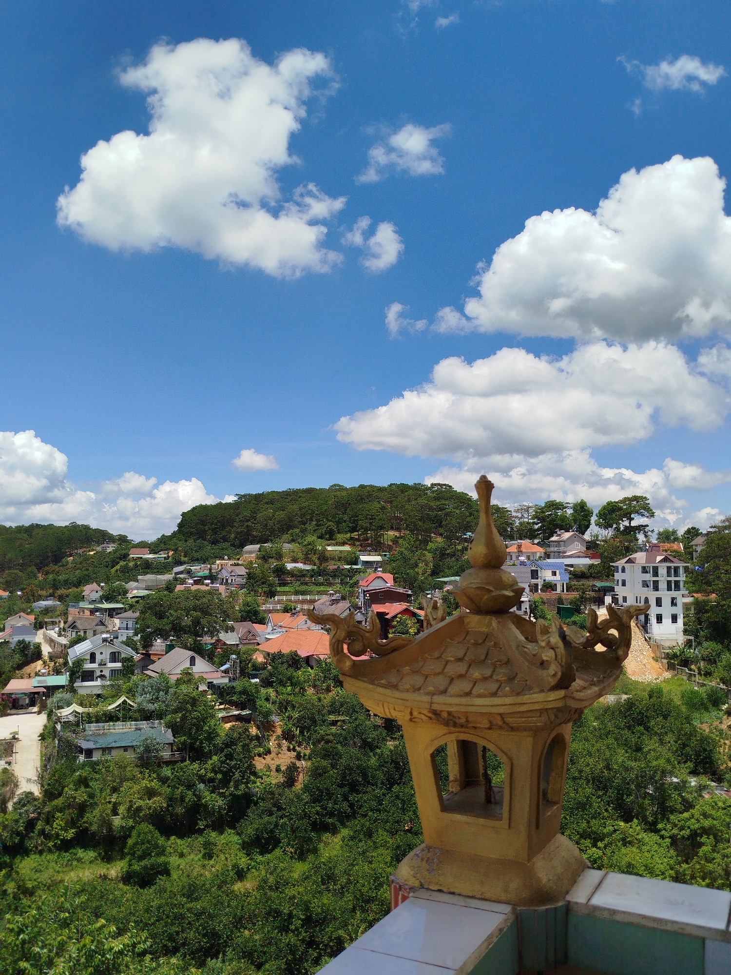 The image shows a daytime view in Da Lat, Vietnam. A golden ornamental lantern stands in the foreground, positioned on a white-tiled surface, possibly a wall or railing. Behind it, a hillside town is visible, composed of various houses with red, white, and grey roofs nestled among trees and green foliage. In the background, a forested hill rises. The sky above is bright blue, dotted with scattered white clouds. No people are visible in the shot.