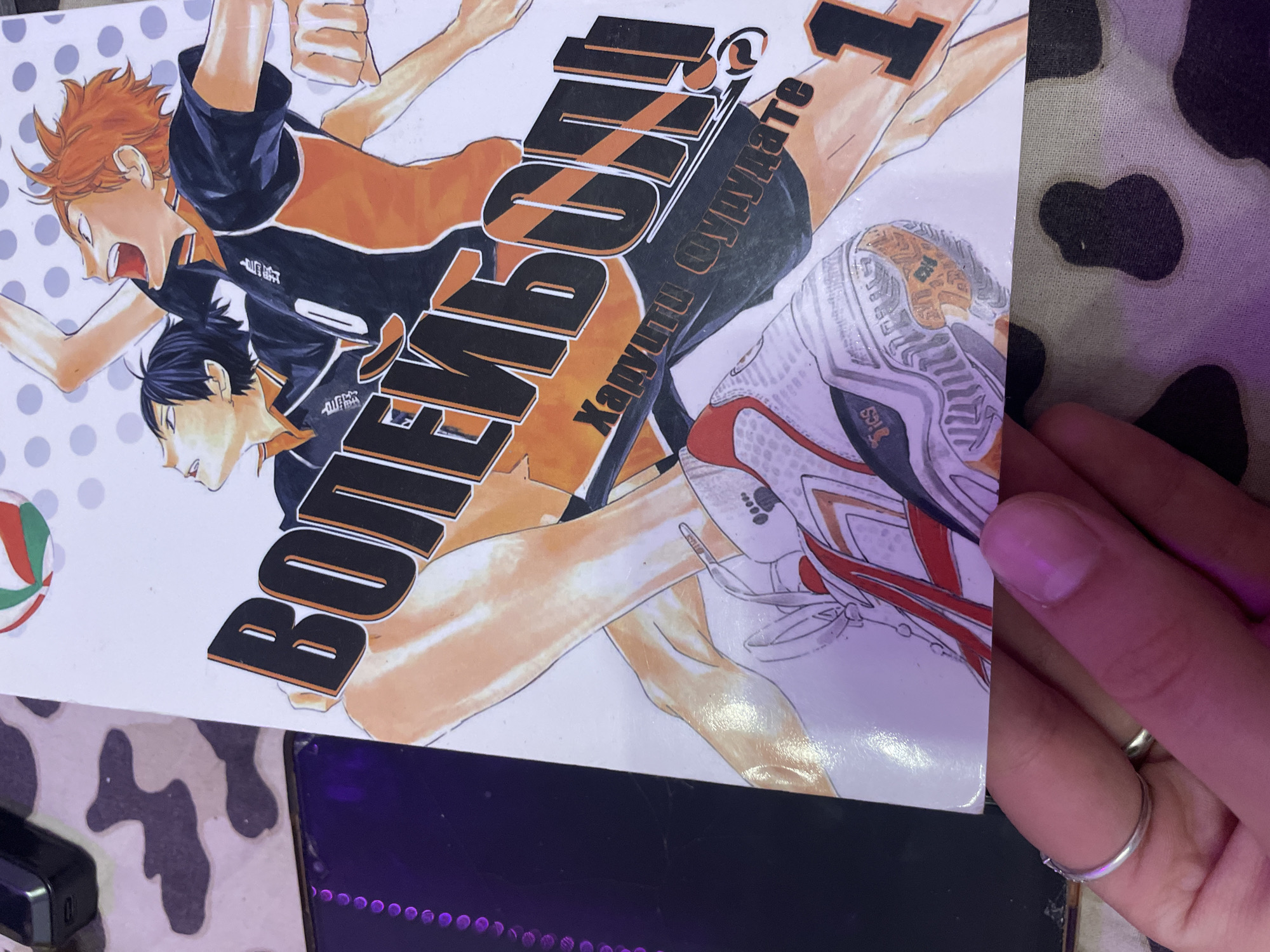 The image shows a close-up of the cover of a manga titled "Haikyuu!!", written in Cyrillic script. The cover features two male anime characters, one with spiky orange hair and the other with black hair, wearing volleyball uniforms. The orange-haired character is reaching upwards, and the black-haired character is looking up. The title "ВОЛЕЙБОЛ!!" is prominently displayed in large, stylized black letters. Below the title, there is smaller text that appears to be in Japanese. The bottom of the cover shows the lower legs and feet of the characters, with one character wearing a white and red volleyball shoe. The manga cover is partially held by a hand with a silver ring on one finger. The background of the image consists of a patterned cloth, possibly a blanket, with black and white cow-like splotches. There are also glimpses of a dark, rectangular object to the left, and some purple lighting is visible on the left side of the image.
There is no information in the image to indicate its location in the city of Irinovka, Russia.