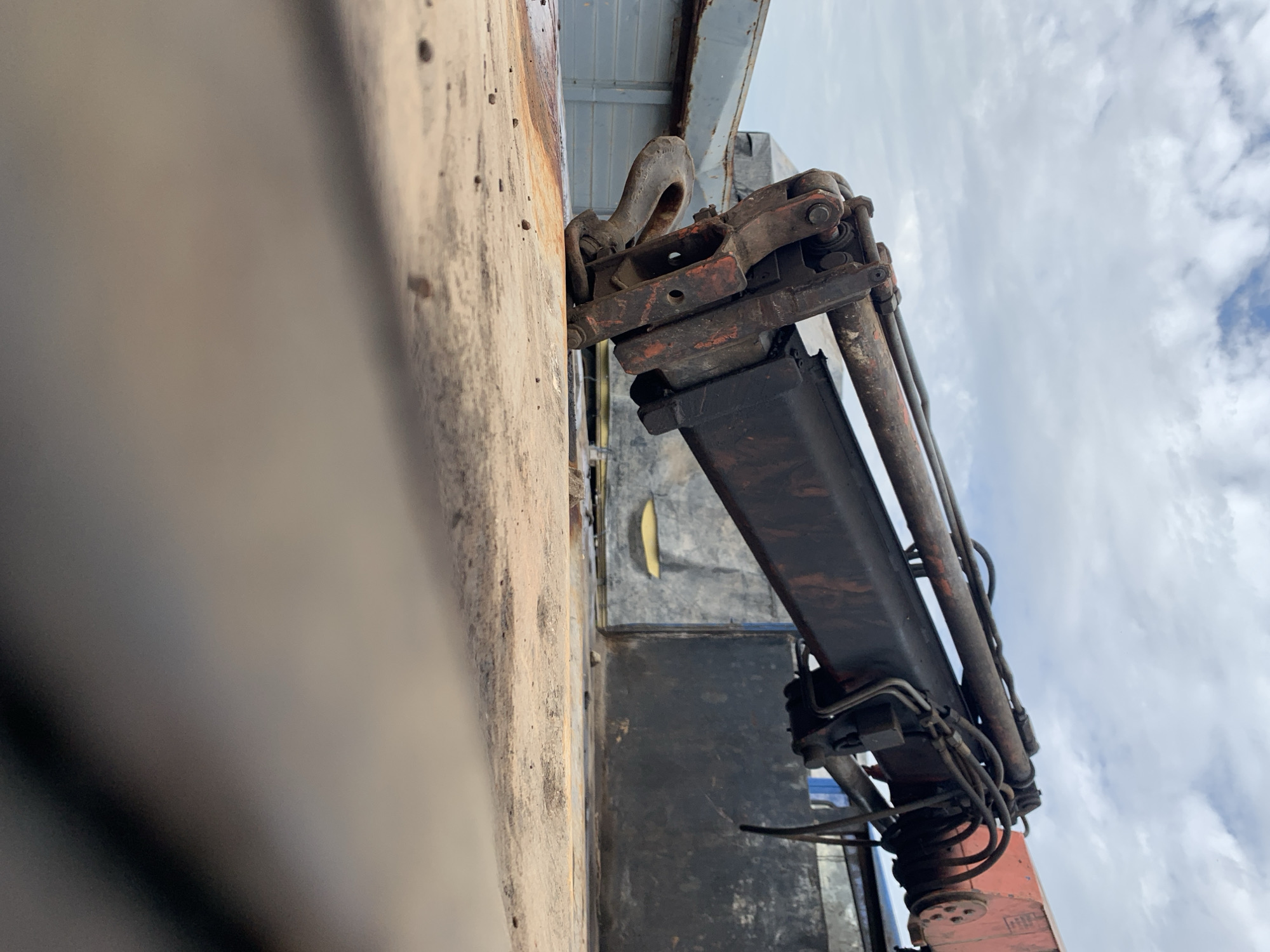 An outdoor, low-angle shot depicts a heavily weathered industrial crane arm and hook positioned over a dirty, flat surface under a cloudy sky in Rafin Par, Nigeria. The foreground consists of a coarse, light brown to tan, possibly wooden, surface scattered with small dark pebbles and debris, appearing as a truck bed or loading platform. Rising from the midground, a robust, dark-colored hydraulic crane arm, exhibiting significant rust and wear with exposed reddish-brown patches, extends diagonally downwards from the upper right toward the lower left. Multiple hydraulic cylinders and black tubing run along the arm. A large, rusty metal hook is attached to the end of the crane arm, resting on or just above the foreground surface. In the background, under a sky filled with white and grey clouds, indistinct industrial structures or vehicle components are visible; a grey metallic structure is present on the left, and a dark, vertical structure on the right. The overall scene suggests an active industrial or transport environment. No individuals are present.