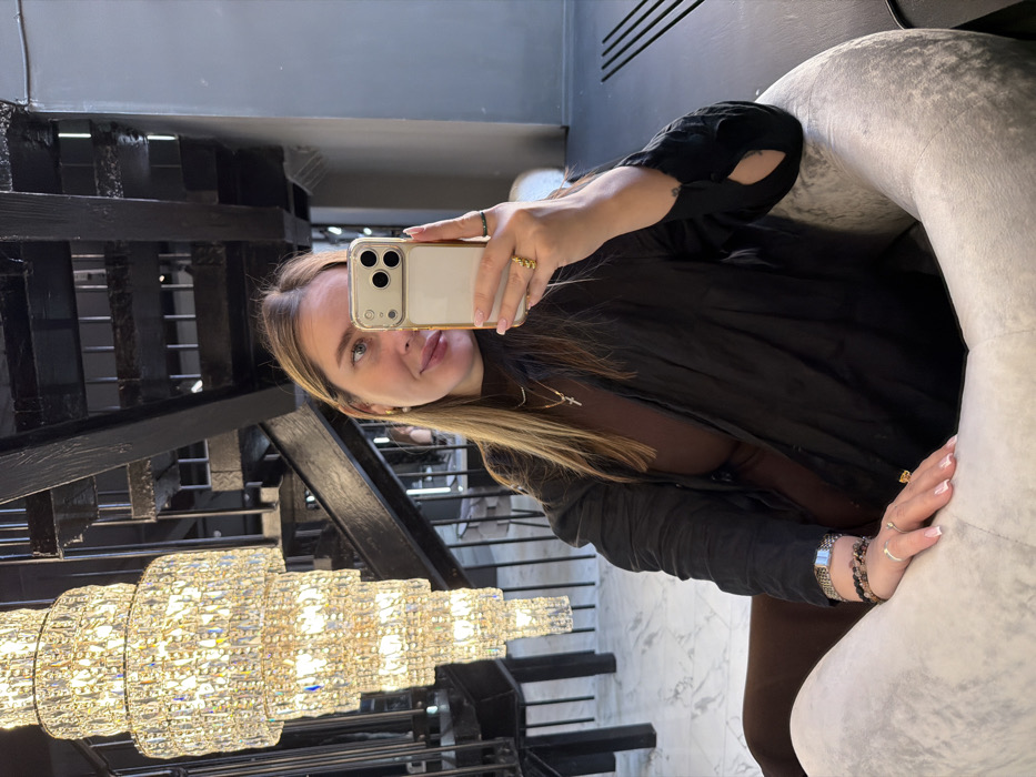 A female subject is shown in a mid-shot taking a selfie within an upscale interior in Moscow, Russia. She has light-toned skin, long light-brown hair, and is wearing a dark brown high-neck top beneath an open black long-sleeved shirt. A gold cross necklace is visible, along with a pearl stud earring on her right ear. Her right hand, holding a light-colored iPhone with a triple-camera system, displays a prominent gold-toned ring on the index finger. Her left wrist features multiple bracelets, including a silver-toned chain and beaded designs, and her nails are manicured with light polish. The subject is seated in a light-gray, rounded armchair, with a dark brown chair partially visible to her left. The background reveals a brightly lit, multi-tiered crystal chandelier on the left and a dark, possibly black, wooden or metal staircase ascending on the right. The flooring consists of white marble with gray veining.