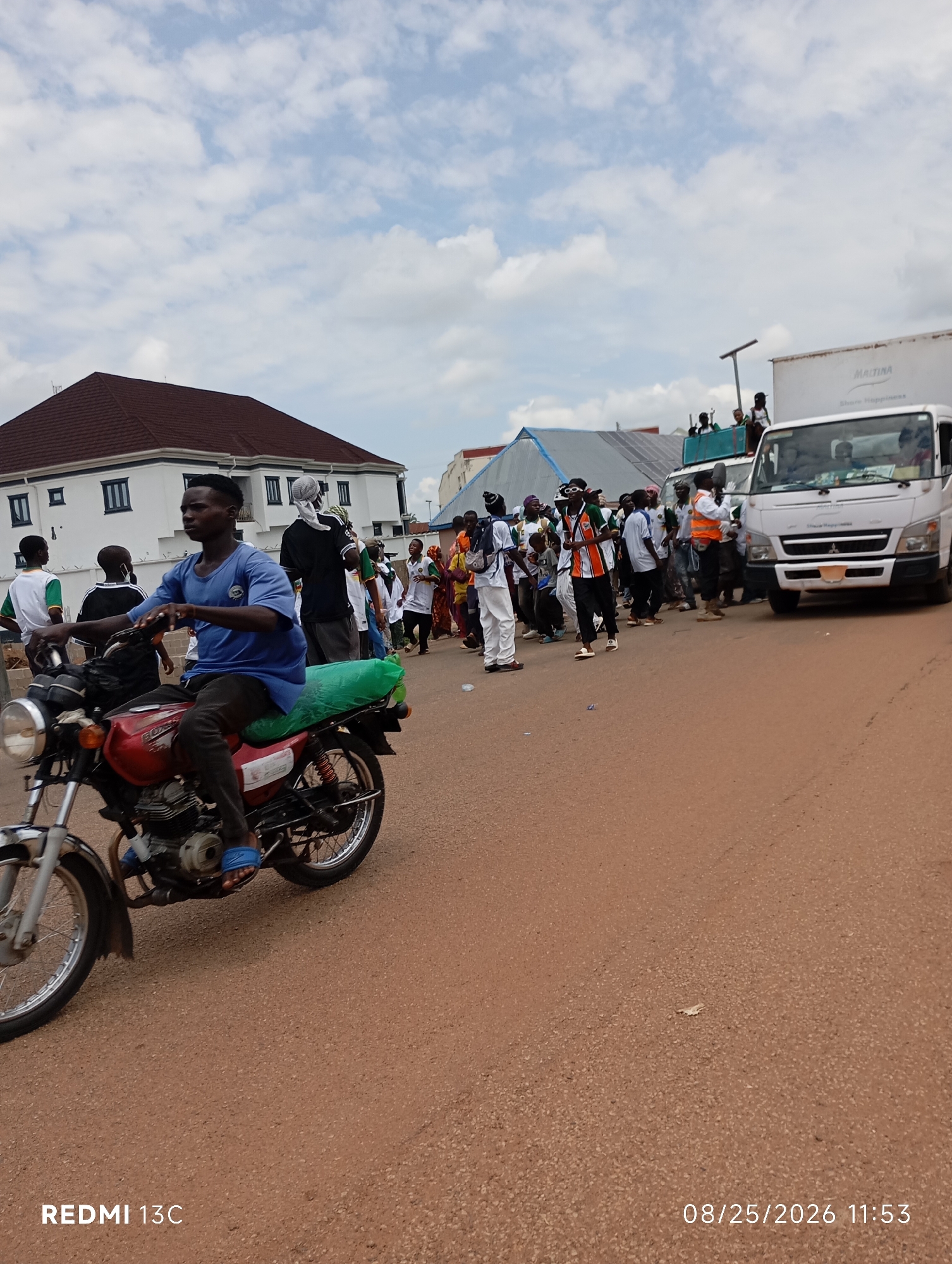 This image depicts a street scene in Shendam, Nigeria, on August 25, 2026, at 11:53 AM. A man is riding a red motorcycle in the foreground, moving from left to right. He is wearing a blue t-shirt and dark pants, with blue sandals. The motorcycle has a green plastic cover on the seat. Behind him, a group of people are walking along the street. Some are wearing white and green attire, while others are in casual clothing. A white truck is parked or moving on the right side of the street. Buildings are visible in the background, including a large white building with a dark red roof on the left. The sky is partly cloudy. The street surface appears to be made of asphalt or a similar material. The overall impression is that of a procession or a gathering of people on a street in Nigeria.