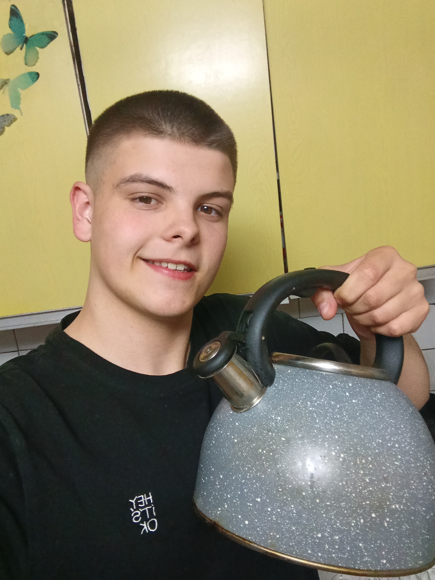 A young man with short brown hair is holding a gray speckled tea kettle with a black handle. He is smiling slightly, and wearing a black t-shirt with reversed white text. The background shows yellow cabinets with a decorative butterfly sticker. The location in the city of Красный Маяк, Russia is not evident in the image.