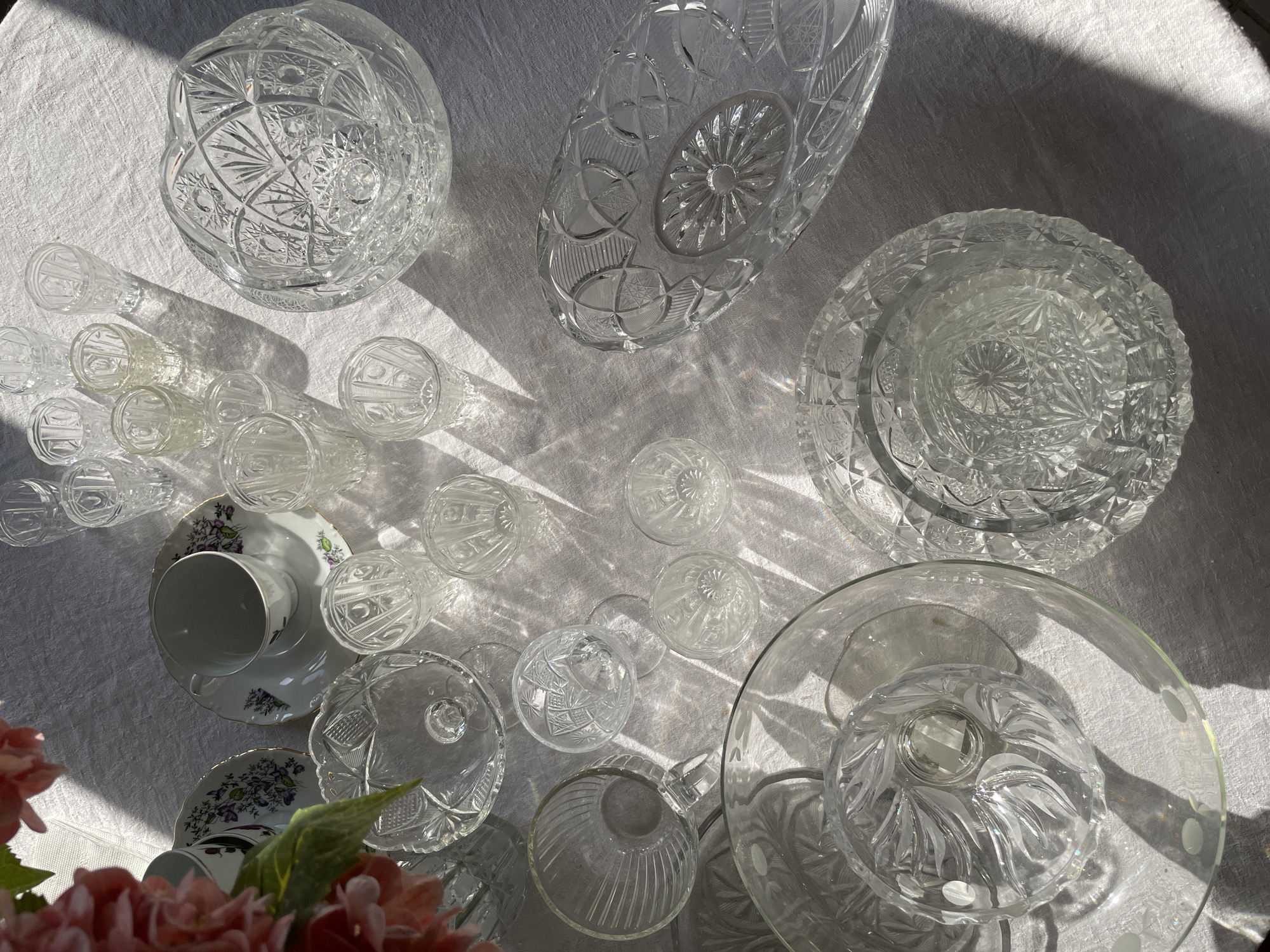 A high-angle shot shows an assortment of crystal glassware and china arranged on a white tablecloth. The light source is strong, casting sharp shadows. The crystal objects include a large bowl, an elongated bowl, a plate, numerous small glasses, and a footed bowl. Two floral-patterned china cups and saucers are also present, partially obscured by a bouquet of pink flowers in the lower left corner. The backdrop is a white, slightly textured fabric. The image was likely taken outdoors or near a large window due to the harsh lighting. No people are visible. The location, Chaikovsky, Russia, is outside the scope of what can be determined from the image itself.
