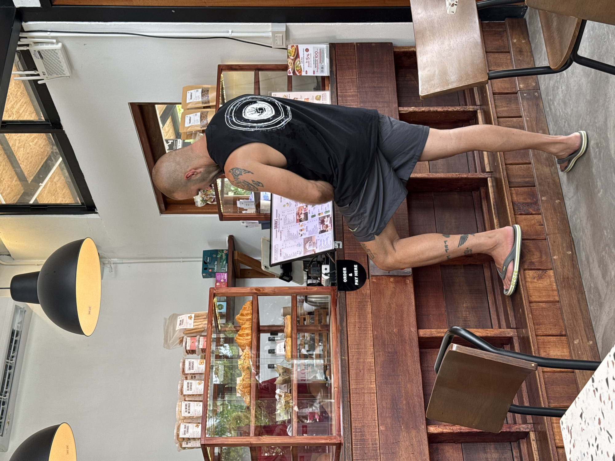 In Ko Samui District, Thailand, a bald man wearing a black sleeveless shirt with a circular graphic and dark grey shorts stands with his right leg propped on a wooden counter. He is facing away from the camera, looking at a digital menu on a tablet. The tablet is situated on the counter, which is made of dark wood. Behind the counter, a glass display case showcases pastries, and shelves above hold packaged cookies and other items. The environment appears to be a cafe or bakery, with bright lighting from overhead fixtures and natural light filtering through a window. A sign on the counter reads "ORDER & PAY HERE." The floor is concrete, and a wooden chair with a black metal frame is visible to the left, with part of a table in the foreground. Tattoos are visible on the man's left arm and right leg.