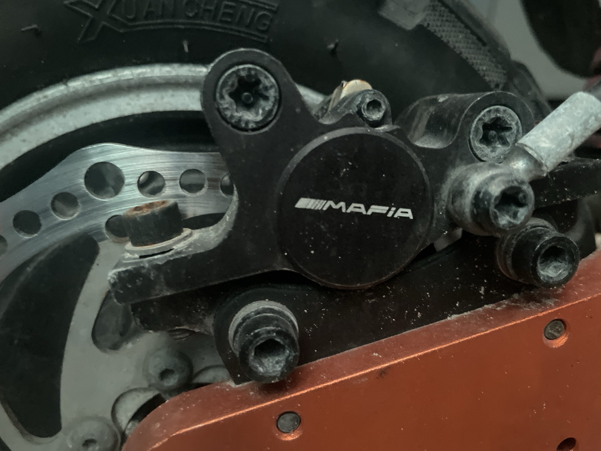 This is a close-up, eye-level shot of a bicycle brake caliper, specifically the disc brake assembly of a scooter or electric bike. The caliper is black and prominently features a logo that reads "MAFIA" in white, stylized text. The brake disc, made of metal with visible holes, is partially obscured by the caliper. Several bolts are visible, some appearing to be hex bolts. The surrounding area shows a black tire with some white lettering that is partially legible as "XUAN CHENG", and an orange component that is likely part of the scooter's frame. The scene is industrial or mechanical, with a focus on the functional components of the vehicle. There is no indication of people, weather, or time of day. The setting appears to be a workshop or storage area.