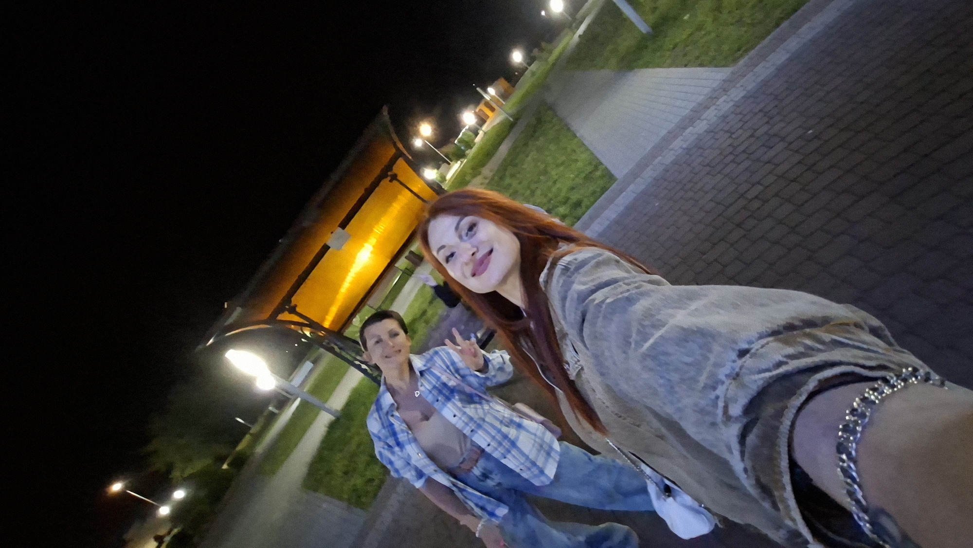 Two women are posing for a selfie at night in a park-like area. The woman in the foreground has long, reddish-brown hair and is wearing a beige jacket and a silver bracelet. The woman behind her has short brown hair and is wearing a blue and white plaid shirt and jeans. She is making a peace sign with her fingers. In the background, there is a covered seating area with orange lighting and streetlights. The ground is paved with bricks. The image appears to have been taken in the city of Myadzyel, Belarus.