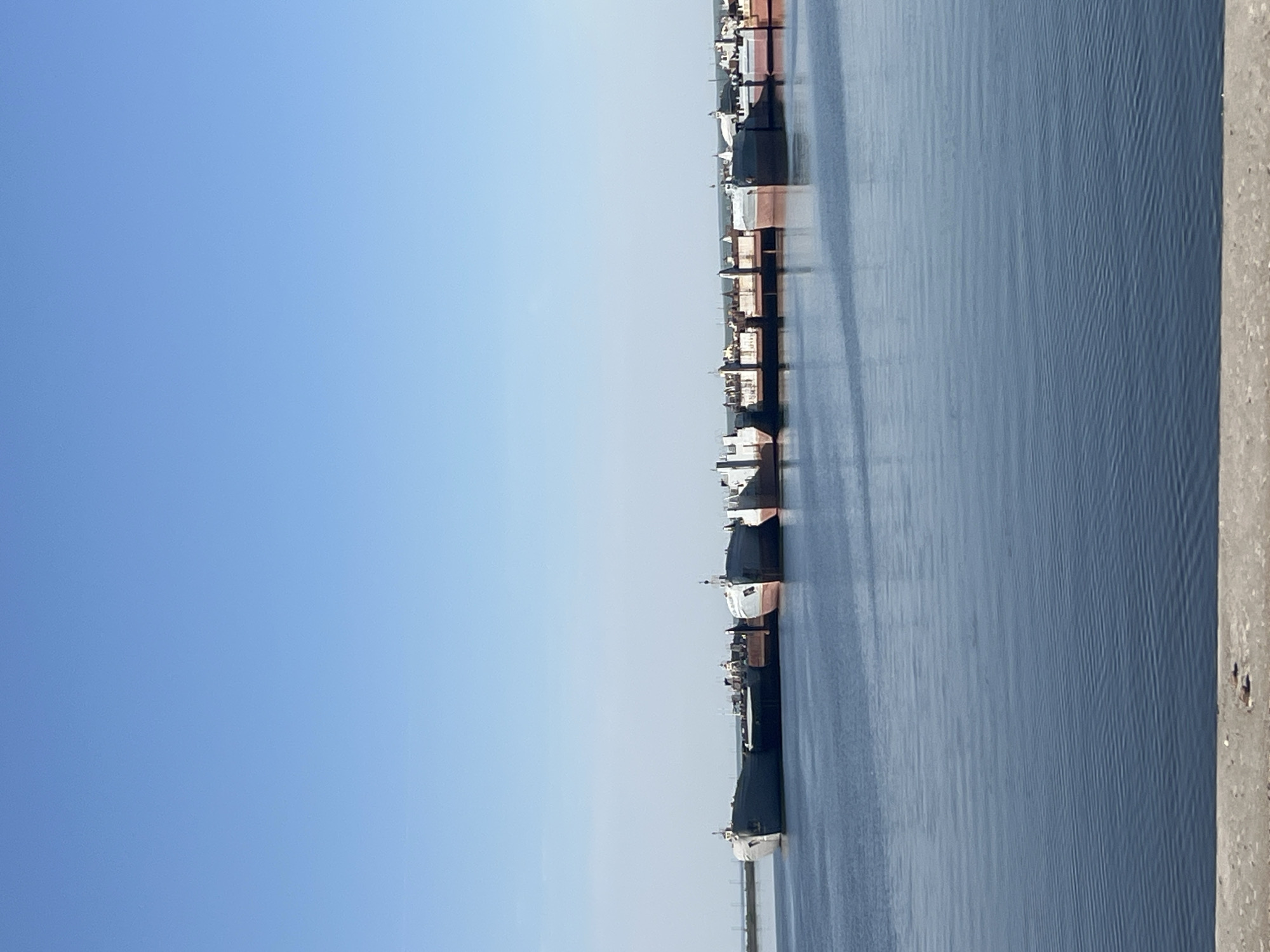 The image shows a body of water with several ships docked along a pier. The sky is clear and blue. The location is Chaikovsky, Russia. The pier is made of wood and concrete. The ships appear to be cargo ships.