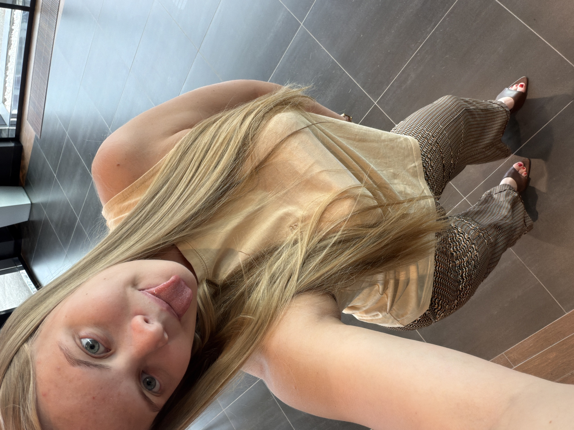 A selfie-style photograph depicts a blonde woman in a light beige shirt and patterned trousers. She is sticking her tongue out towards the camera. She is wearing brown open-toed shoes. The background appears to be a tiled floor, possibly indoors. The location in the city of Izhevsk, Russia is not determinable from the image.