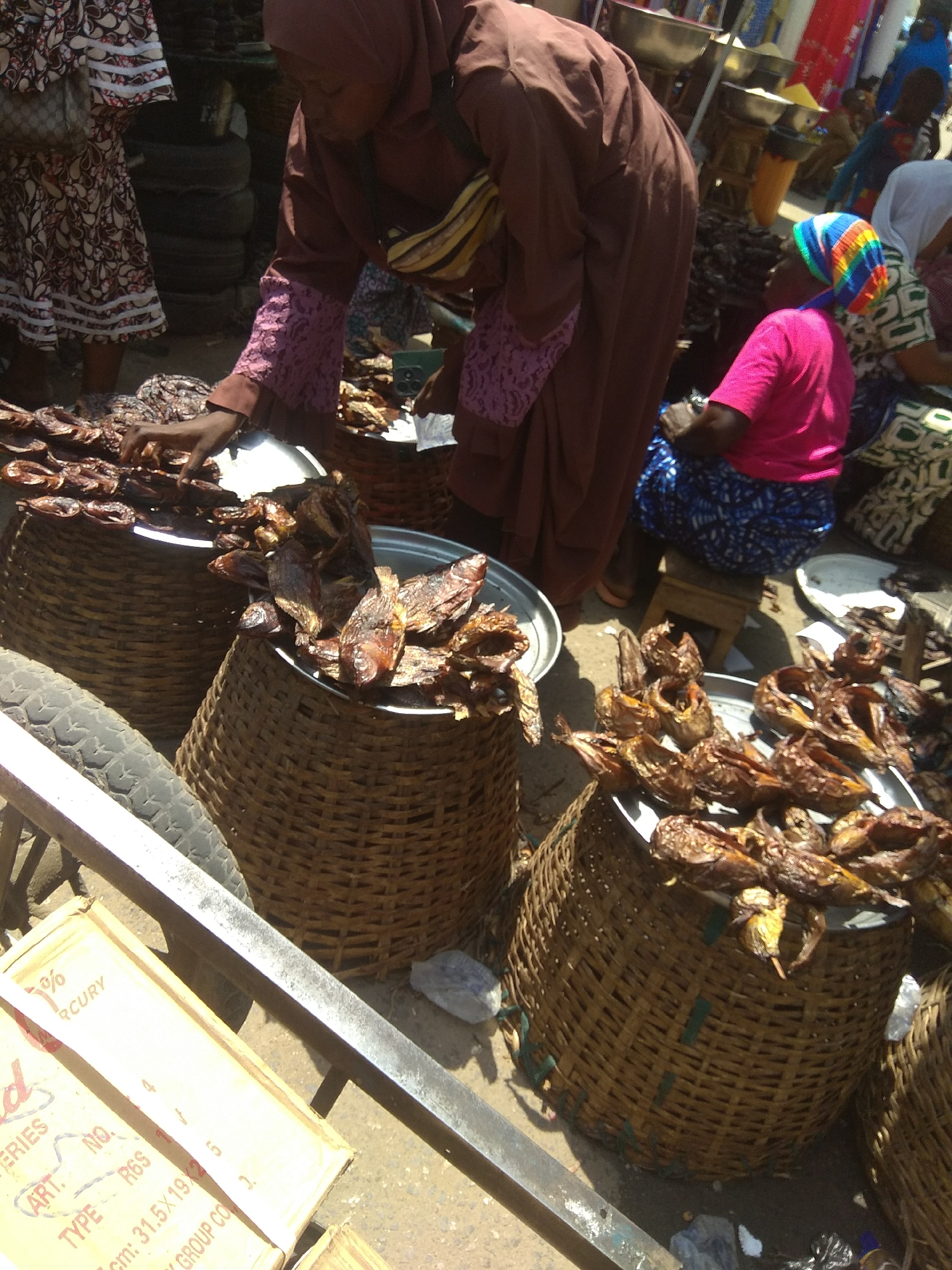 This media file captures a vibrant scene at a market in Jalingo, Nigeria, likely during daylight hours given the strong shadows. The central focus is on a woman in a dark brown hijab and matching robe, with striking purple lace on her sleeves. She is reaching into a large woven basket filled with dried fish. Her attire suggests a conservative cultural context.
Beside her, another woman, also wearing a hijab, is seated on a small stool. She has a brightly colored, striped headscarf and is wearing a vibrant pink t-shirt, contrasting with the more subdued colors of the woman reaching into the basket. Another woman, partially visible, wears a white hijab and a patterned garment. The market setting is evident from the array of woven baskets filled with produce, specifically dried fish. The baskets are large and sturdy, typical of market stalls.
In the foreground, a cart with a tire and metal railing is visible, suggesting the movement of goods. Several boxes are stacked on the cart, one of which has visible text. The text on the box includes "RCURY," "ERIES," "ART. NO.", "TYPE R6S", and dimensions "31.5×19×2.5 cm," and "GROUP CO." This indicates some of the products being sold or transported in the market. The overall atmosphere appears busy and active, a common characteristic of Nigerian markets. The image is taken from a slightly elevated angle, looking down on the scene.