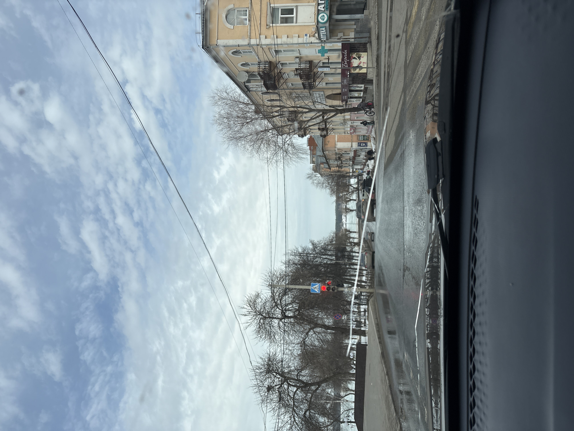 The image shows a view from inside a car driving along a wet street in Votkinsk, Russia. The sky is cloudy. On the left side of the road are bare trees and beyond them, a body of water. On the right side is a multi-story building with a pharmacy ("АПТЕКА" in Russian) sign. A pedestrian crossing with a traffic light is visible further down the road, showing a red signal. A cyclist is on the sidewalk near the pharmacy building. Other buildings are visible further down the street.