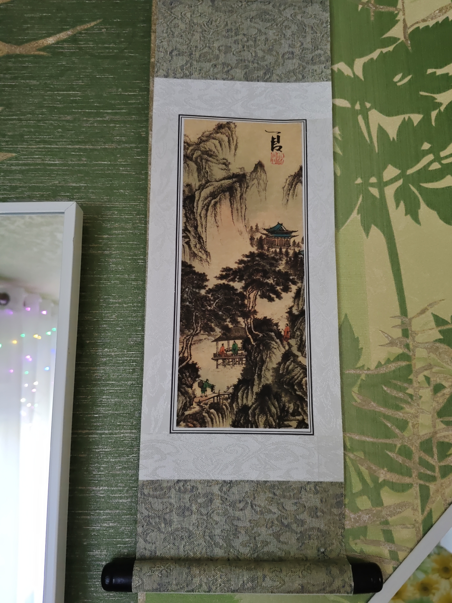 The image shows a scroll painting of a traditional Chinese landscape. The painting depicts a mountainous scene with a pagoda-like building nestled among trees and rocks. Two figures are visible in the foreground, one walking with a pole over his shoulder and another standing near a bridge. The scroll is hung on a wall that has green textured wallpaper with a bamboo motif. To the left of the scroll, a white-framed mirror is visible, reflecting some colored lights and what appears to be part of a room's interior. The painting is framed by white and patterned fabric borders. The bottom of the scroll is rolled around a dark cylindrical rod. The scene is indoors, likely in a residential setting. There is no visible indication of the location being in Rostov-on-Don, Russia.