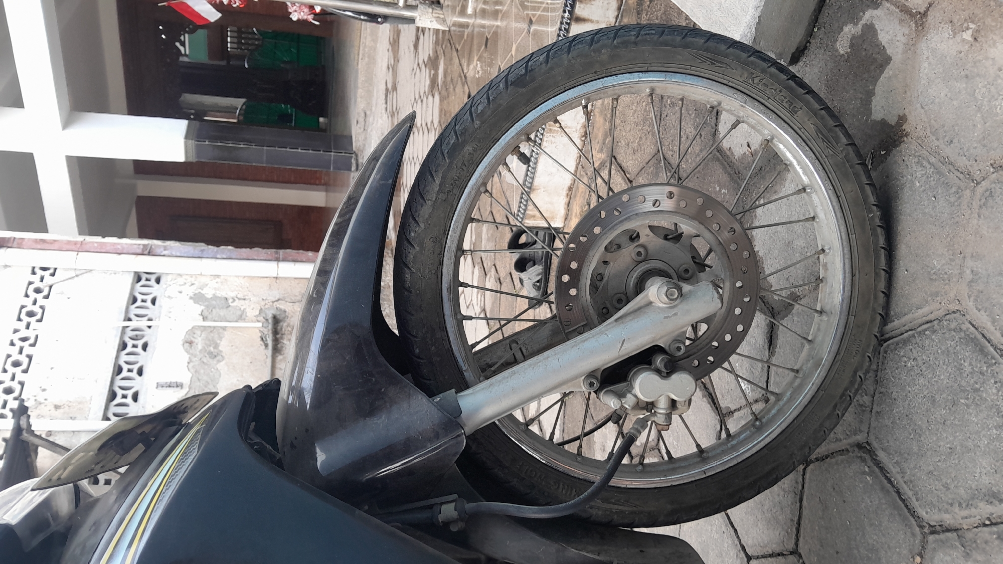 The image shows the front wheel and fender of a dark-colored motorcycle parked on a paved surface, likely in Sambong, Indonesia. The motorcycle is positioned in front of a building with white decorative grilles and a tiled roof. In the background, an Indonesian flag can be seen flying from a pole. The ground appears wet in places, suggesting recent rain. A pair of black sandals are visible on the ground behind the wheel. The scene is outdoors and appears to be daytime.