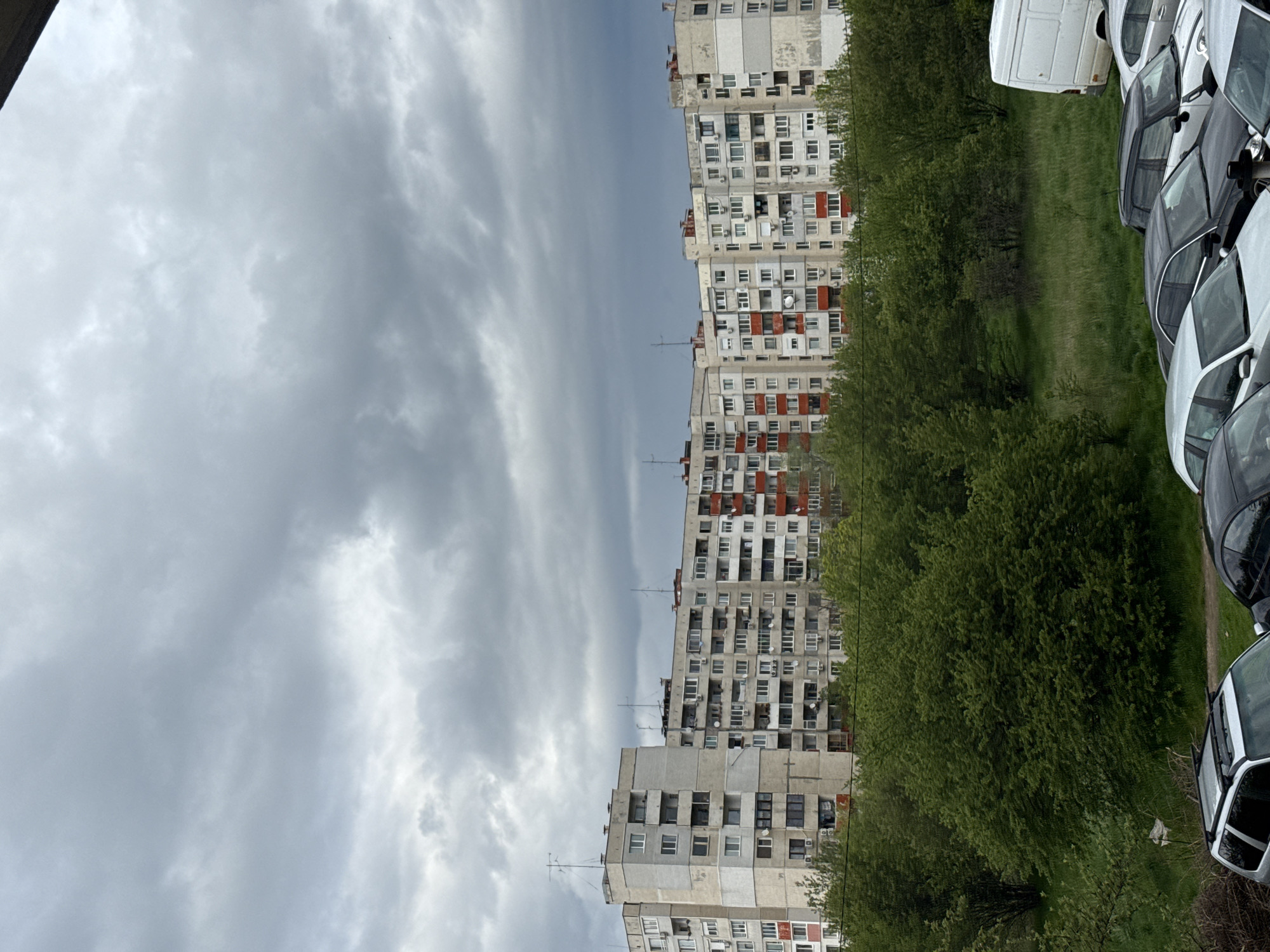 The photograph depicts an urban residential scene dominated by several multi-story apartment buildings under a cloudy sky. The buildings, visible on the left side of the frame, are constructed from concrete, featuring numerous windows, balconies, and external air conditioning units. Some sections of the buildings appear weathered, and a few balconies are accented with red paint, providing a contrast against the predominantly gray and off-white facades. The architecture is characteristic of Eastern European post-socialist urban planning.
A strip of vibrant green trees and grass runs horizontally across the middle of the image, acting as a natural divider between the apartment blocks and the right side of the frame. On the far right, a line of parked vehicles is visible, including a white van and several cars of different models and colors (e.g., black, white).
The sky above is heavily overcast, filled with various shades of gray clouds, indicating a cloudy or potentially rainy daytime. There are no people visible, and no discernible activity or event is taking place, presenting a static, everyday view of a residential area. No visible text is present in the image. The overall impression is one of typical urban life in a city like Sofia, Bulgaria.