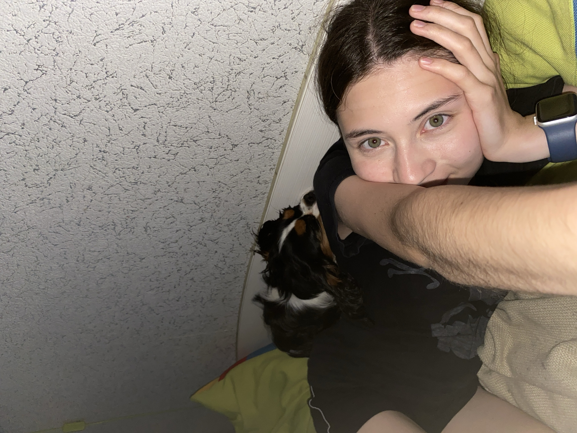 A tri-color Cavalier King Charles Spaniel lies on the chest of a person who is lying on their back, propped up on pillows. The person is looking at the camera with their left hand supporting their head. A dark-colored smartwatch is visible on the person's left wrist. The background consists of a textured wall and a white baseboard. The image appears to have been taken indoors in a residential setting.