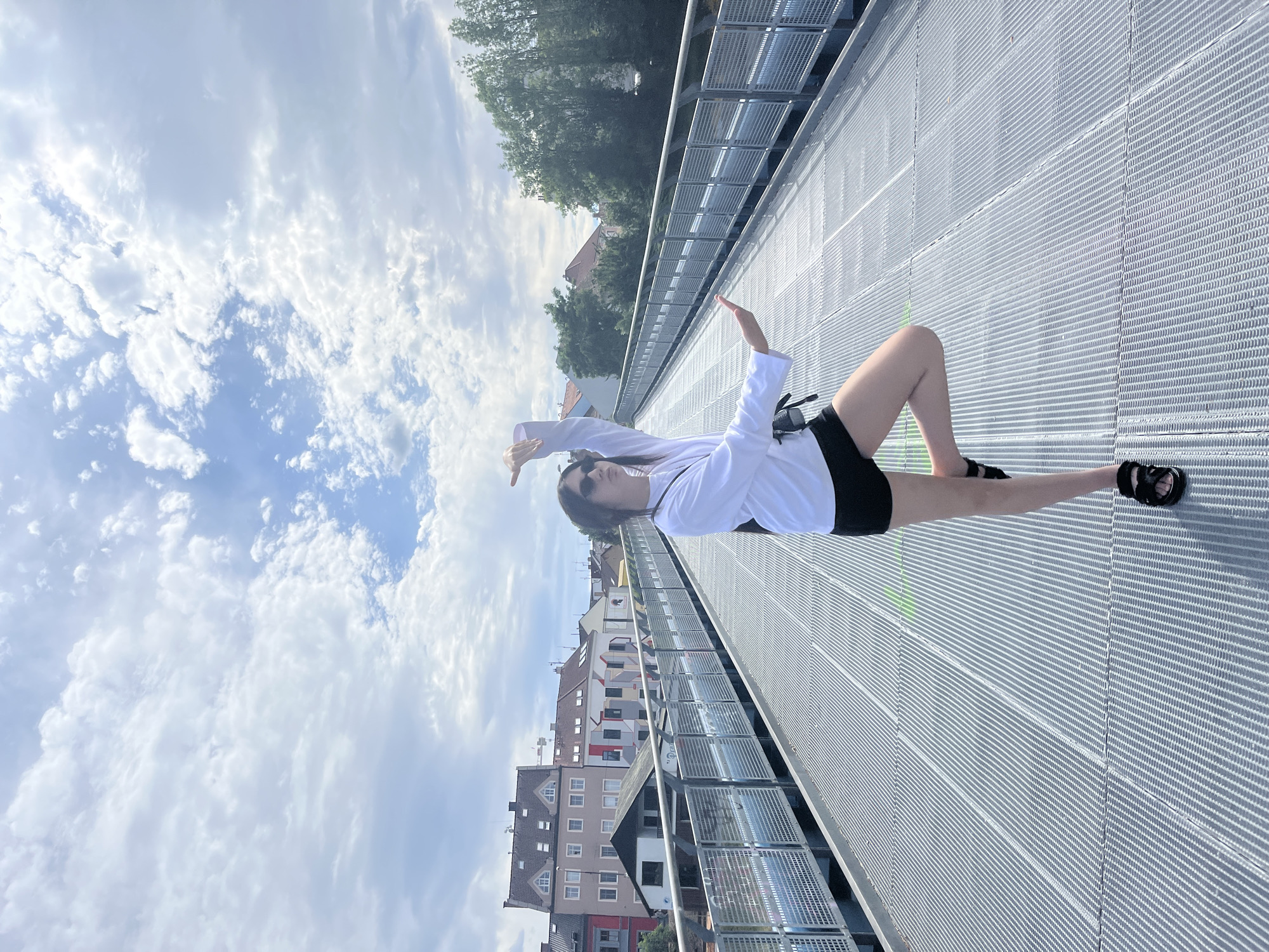 A young woman stands on a modern metal pedestrian bridge in Pilsen, Czechia, balancing on one leg in a dance-like pose. She is wearing a white long-sleeved shirt, black shorts, black sandals, and sunglasses. The bridge, with its metal grate surface and railings, spans over a body of water, though the water is not clearly visible. In the background, buildings with varied architectural styles line the riverbank, characteristic of Pilsen's urban landscape. Trees and a cloudy sky complete the scene, suggesting it is daytime. The low angle of the sun indicates it might be morning or afternoon. The bridge appears to be a recent addition to the city's infrastructure.