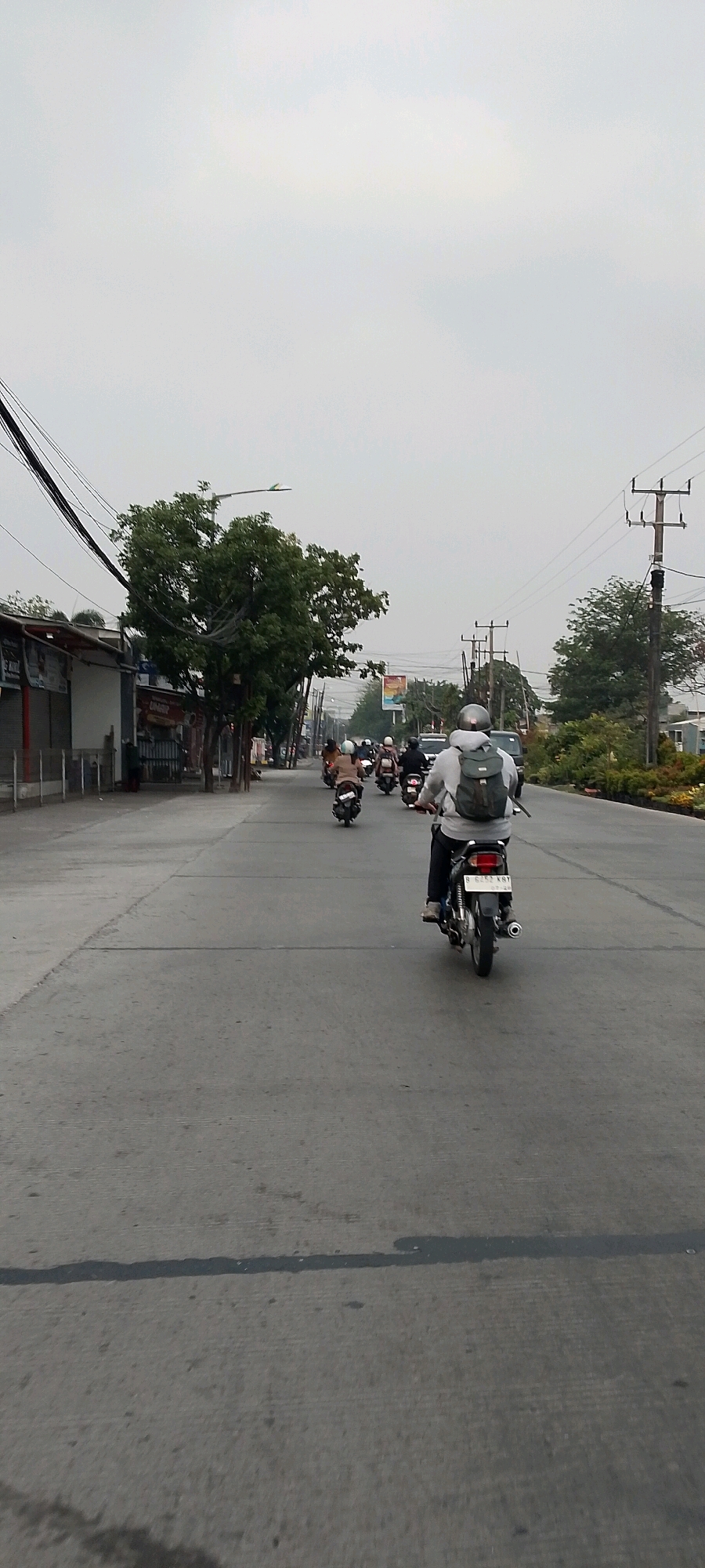 The image depicts a street scene in Bekasi, Indonesia, on a cloudy day. The foreground is dominated by a paved road with visible lane markings. A person wearing a gray hoodie and a backpack rides a motorcycle forward, away from the camera. The motorcycle has a license plate that reads "B 6252 XRY". In the middle ground, several other motorcycles are also traveling in the same direction. On the left side of the road, there are buildings and trees lining the street, with power lines overhead. On the right side, more trees and vegetation are visible, along with utility poles and more power lines. The overall atmosphere is that of an urban environment with moderate traffic.