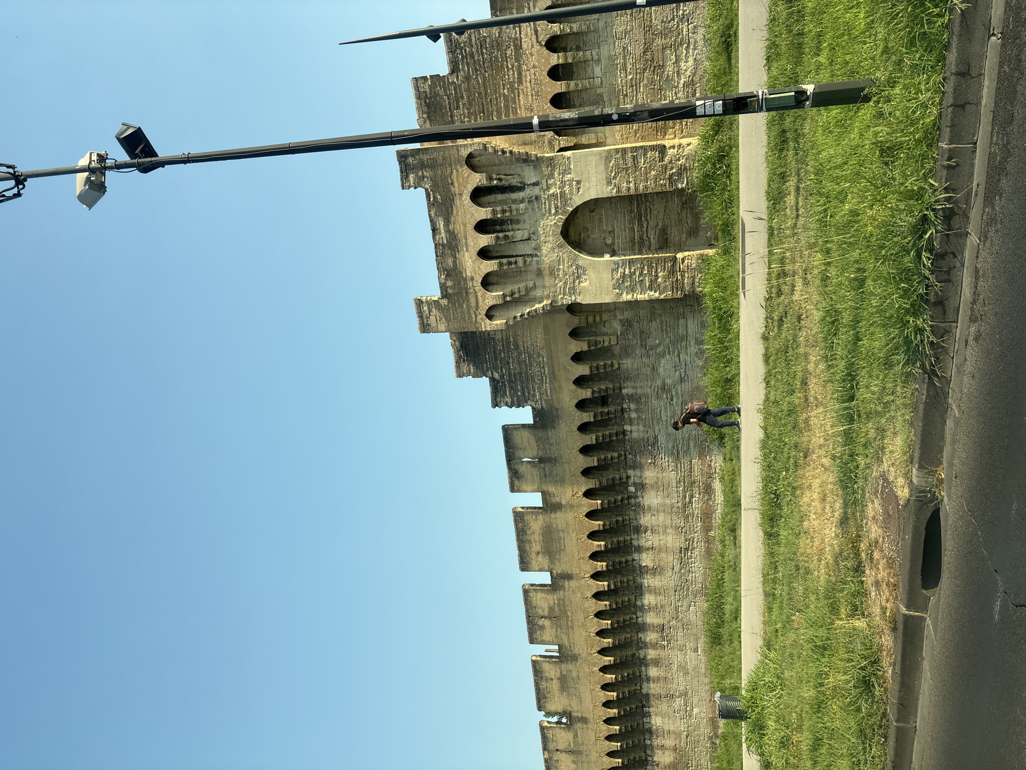 A man with a backpack walks along a paved path next to a section of the medieval ramparts of Avignon, France. The ramparts, constructed of stone, feature crenellations and arched openings. A modern lamppost with attached devices stands to the right of the frame. The ground is covered in green grass, and a paved road is visible in the foreground. The sky is clear and blue.