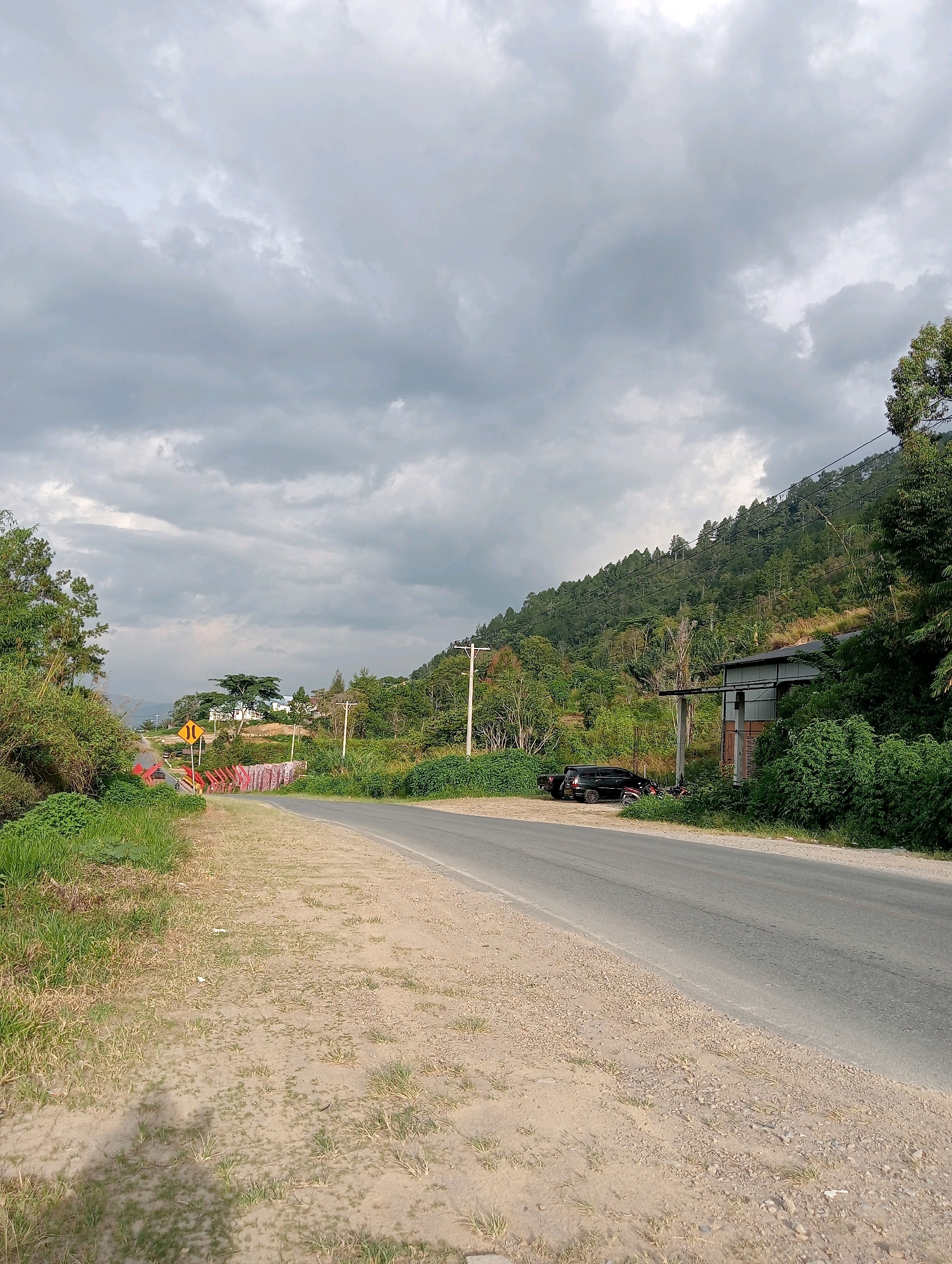The image depicts a roadside scene in Hinalangbagasan, Indonesia. A paved road curves to the right, with a dirt and gravel shoulder on the left. Lush green vegetation lines the roadside, with bamboo and other trees on the left and denser foliage and trees on the right. In the background, a steep, tree-covered hill rises. A few power poles are visible along the road.
On the right side of the road, a simple, utilitarian building with a corrugated metal roof and weathered walls is situated at the base of the hill. In front of the building, a black SUV and a few motorcycles are parked.
Further down the road on the left, a yellow traffic sign with a black arrow indicating a curve is visible. A red railing and some colorful fabric are also present near the roadside. The sky is heavily overcast with dark, cloudy formations. The overall impression is a rural or semi-rural environment with a cloudy, possibly impending weather condition.