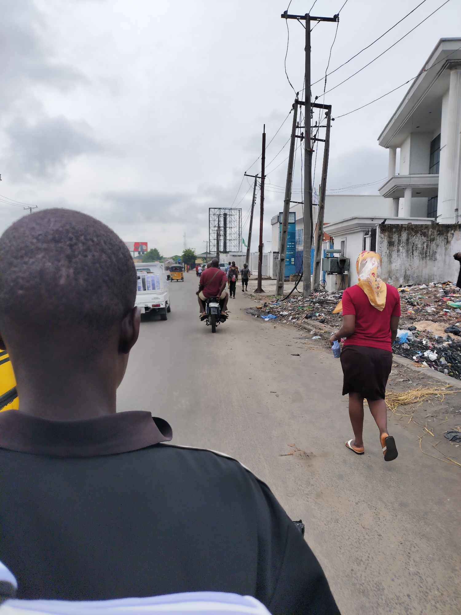 The image presents a street scene in Lagos, Nigeria, viewed from the perspective of someone in the immediate foreground, whose back of the head and shoulders, clad in a black collared shirt, are visible. The sky is overcast, suggesting a cloudy day.
The paved road, showing signs of wear, extends into the distance. Various people and vehicles are moving away from the camera. In the midground, a man rides a motorcycle, wearing a reddish-brown top. Ahead of him, a white mini-truck, possibly branded "KAROO," carries multiple large bottles of water. Further down the road, an auto-rickshaw and other pedestrians can be seen.
Along the right side of the road, a substantial and untidy pile of mixed garbage and debris, including plastics and other waste, lines the street. A woman, wearing a red shirt, a dark skirt, and flip-flops, with a patterned headscarf, walks along the edge of this refuse, holding a light blue bag.
The left side of the street features several tall electrical poles with numerous overhead power lines. Buildings of varying styles are visible on both sides; modern-looking white structures with architectural details are prominent on the right, where a blue signboard with partially visible text "1 800 00" and "ZINASAV" is affixed. In the background, a large, empty metal billboard structure rises against the cloudy horizon. The scene depicts a bustling urban environment grappling with infrastructure and waste management challenges.