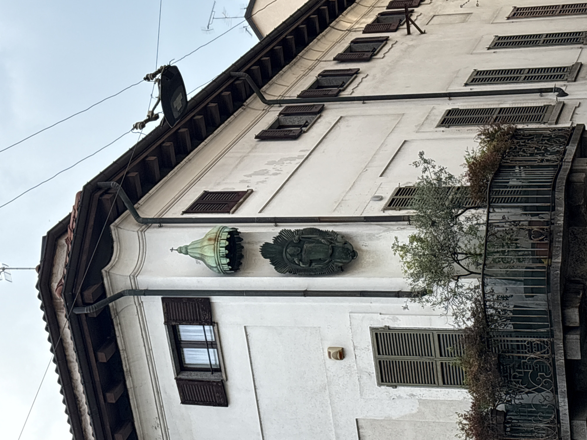 The image shows the exterior of a building in Milan, Italy. The building has a white facade, brown wooden shutters on the windows, and a dark roof. A wrought-iron balcony extends from the building. Ornate metal decorations adorn the wall, including a verdigris-colored dome-shaped object and a dark, round plaque featuring figures. Utility lines are visible in the sky.