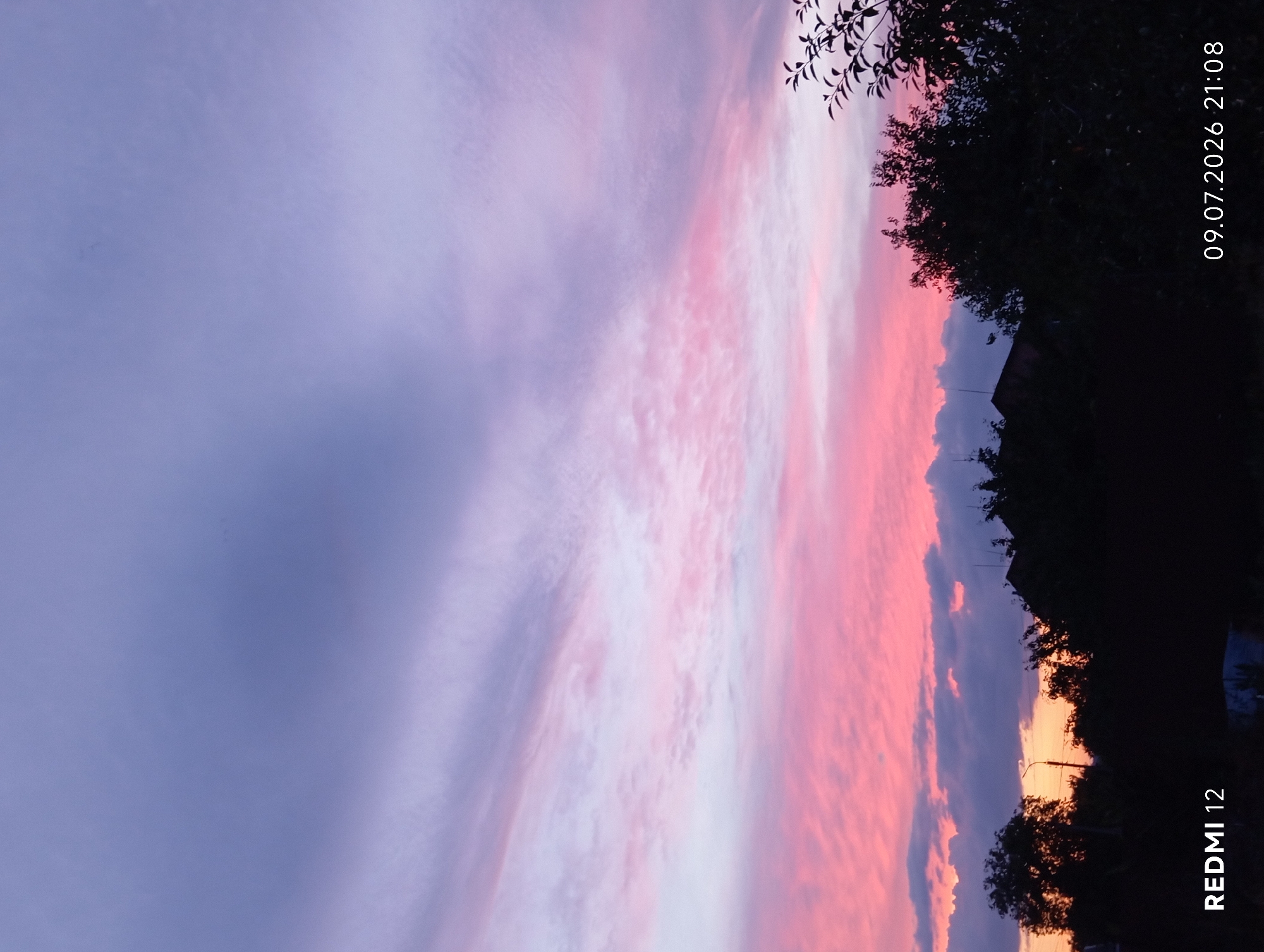 This is a vertical, full-frame photograph capturing a sunset over a silhouetted landscape. The sky is filled with a dramatic gradient of colors, transitioning from a deep purple at the top to vibrant pinks and lighter purples lower down. Wispy clouds are scattered throughout, catching the fading sunlight. In the foreground, the silhouettes of trees and rooftops are visible against the colorful sky. The bottom left corner of the image displays the text "REDMI 12", and the bottom right corner shows the date "09.07.2026" and time "21:08". The scene suggests an evening in a residential area, likely a suburb or outskirts, with the elements of nature and human habitation merging as day turns into night.